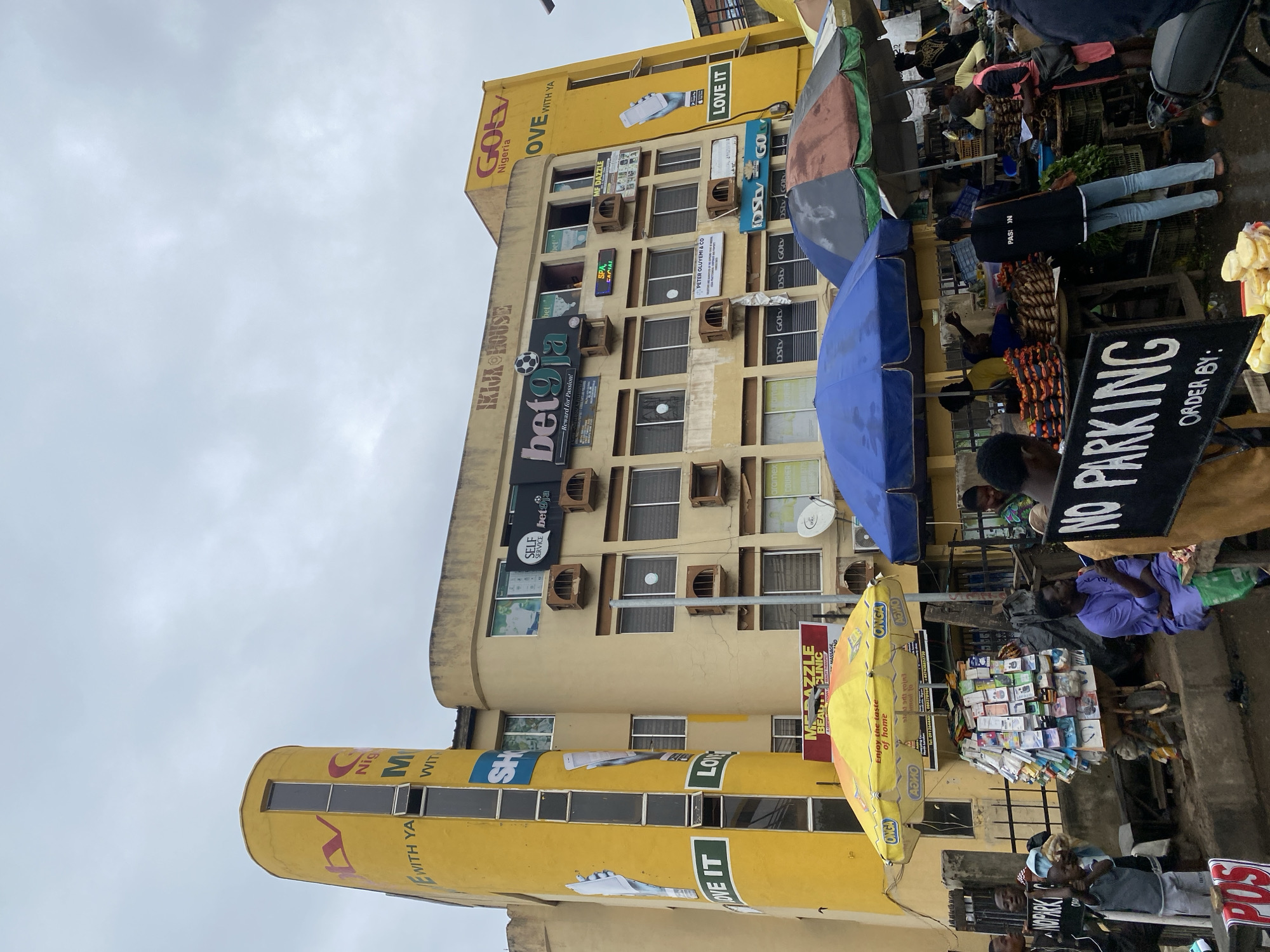 The image shows a street scene in Abeokuta, Nigeria, featuring a bright yellow building with curved architectural elements. The building displays various signs, including "IKIJIA HOUSE," "GOtv Nigeria," "bet9ja," and "LOVE IT." Several windows are visible, some with air conditioning units mounted outside. In the foreground, there are market stalls set up under umbrellas. A cart laden with small items is positioned near the center, and people are browsing the goods. Two "NO PARKING" signs are also visible, one partially obscured. The overall impression is of a busy urban marketplace.