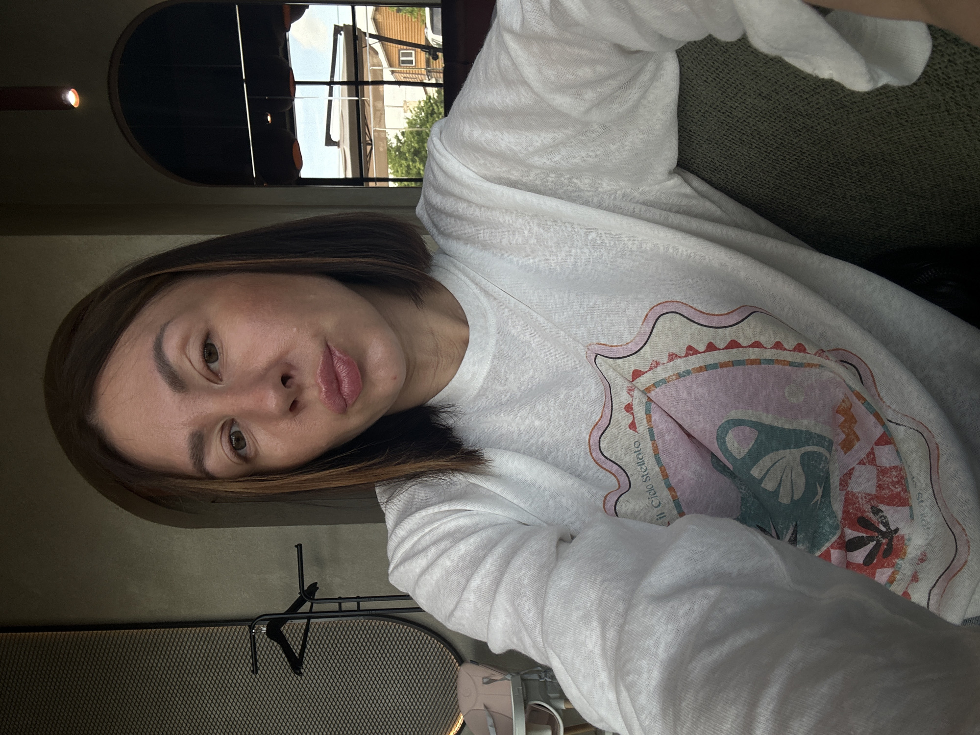 A woman is depicted in a close-up selfie, centrally framed, seated indoors in what appears to be a cafe or restaurant in Усады, Russia. She has medium-brown, bob-cut hair with subtle lighter streaks visible, particularly on the left side of the frame. Her eyes are open, with dark irises. Her lips are pursed in a pouting expression. She is wearing a white long-sleeved top featuring a multi-colored graphic design on the chest, which includes abstract shapes and the partial text "il Cielo Stellato." The fabric of the top has a textured, slightly sheer appearance.
The indoor background features walls with a dark greenish-grey, textured finish. Two arched openings are visible: one to the left and one to the right of the woman. To the left, behind her, a dark metal grid-like panel and a black garment rack are visible. Further left and slightly behind, a light pink baby high chair is partially visible. To the right, through the archway, a multi-paned window provides a view of an outdoor scene. A red cylindrical light fixture emitting a warm glow hangs from the ceiling above the right arch. The woman is seated on dark green upholstered furniture, visible in the lower right portion of the frame.
The outdoor view through the window shows a bright sky, green trees, and multiple structures. A light-colored awning or umbrella structure is prominent, along with a building or house-like structure with a dark roof and a white-framed window. A portion of a white vehicle is also visible on the far right of the outdoor scene.
