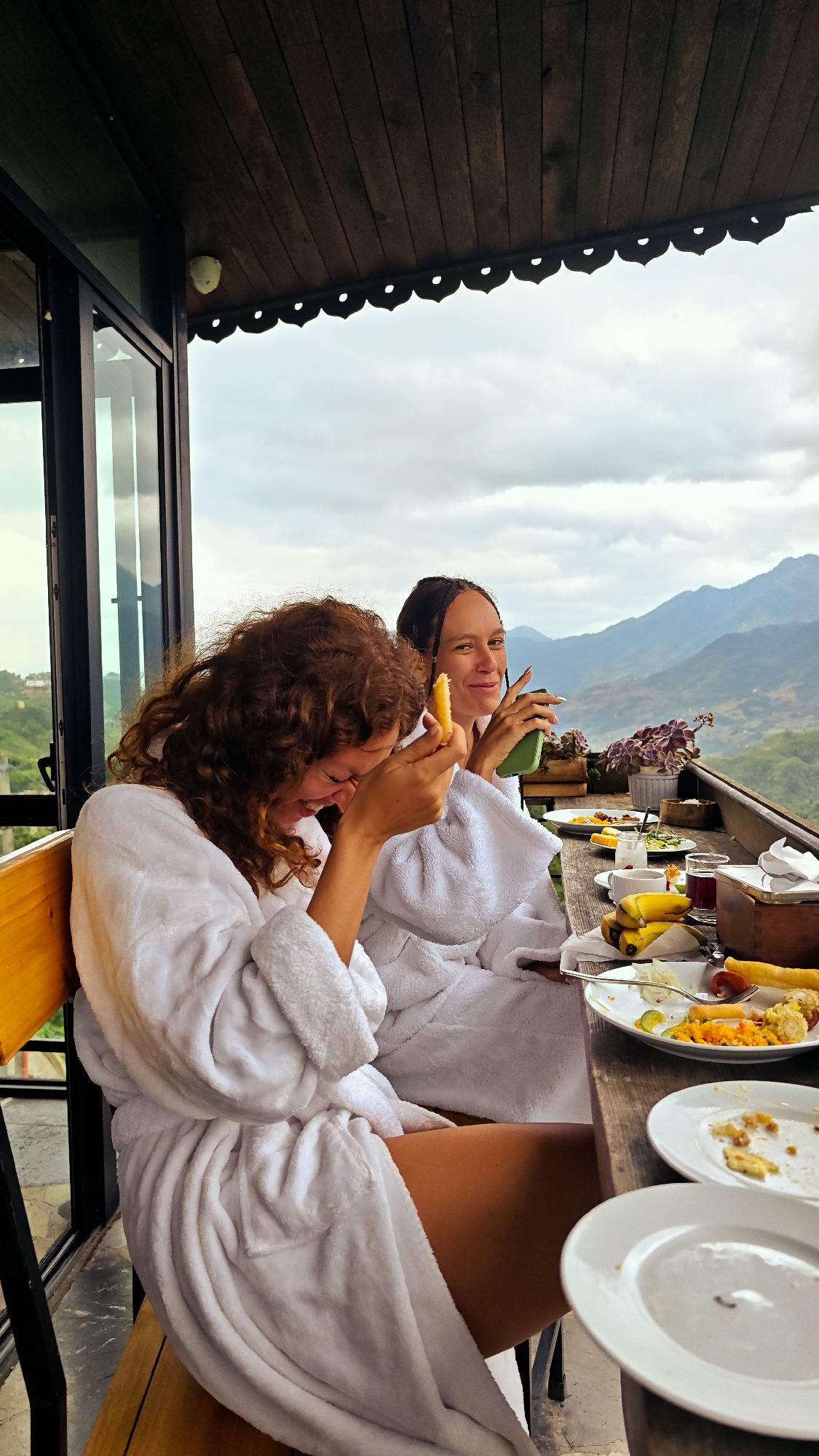 Two women wearing white robes are seated at a wooden table on a balcony. The woman on the left has curly auburn hair and is laughing, holding a yellow object near her face. The woman on the right has dark hair pulled back, a slight smile, and is holding a green drink. The table is cluttered with plates, food remnants, bananas, and drinking glasses. Behind them, a view of mountains and foliage is visible under a cloudy sky. The balcony is enclosed by a glass window and black metal frame on the left and a wooden railing on the right. The location appears to be in Sa Pa Ward, Vietnam, as evidenced by the mountainous landscape and the architectural style of the structure.