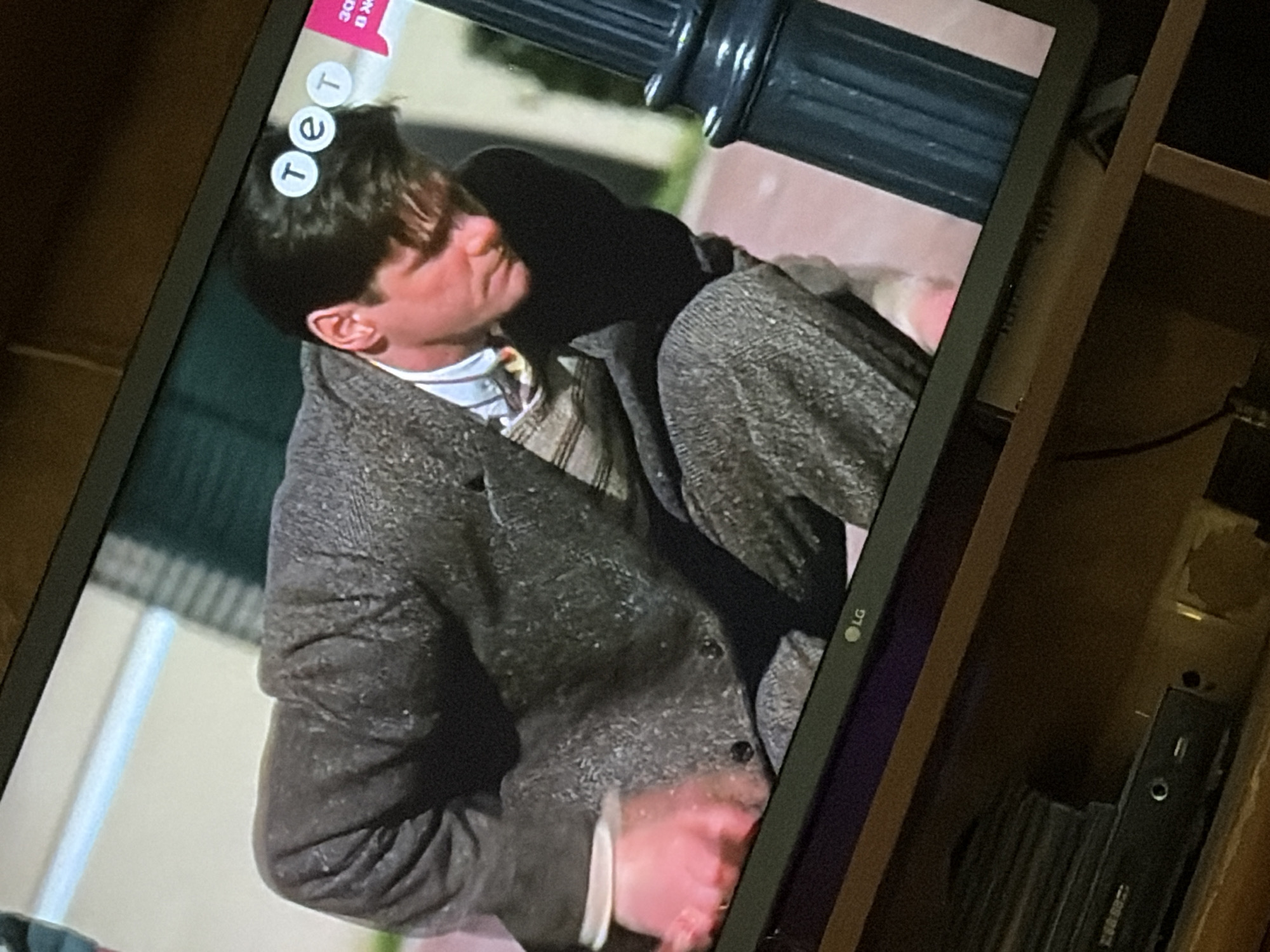 A television screen displays a scene from a movie or show. In the foreground, a man with dark hair is hunched over, wearing a grey tweed suit over a collared shirt and a patterned sweater vest. His right hand, illuminated with a pinkish hue, is extended downwards, possibly touching something on the ground. The background of the screen shows a blurry outdoor setting with a light-colored wall and green foliage. In the upper right corner of the screen, three white circles with blue lettering are visible, forming the logo "TET," a Ukrainian television channel. The LG logo is present in the bottom left corner of the television screen. The television itself is placed on a wooden shelf, with electronic devices and black discs visible on the shelf below. The overall scene is captured from a slightly elevated angle, showing the television screen and its immediate surroundings. The image does not provide information about the specific location within the city of Osolynka, Ukraine.