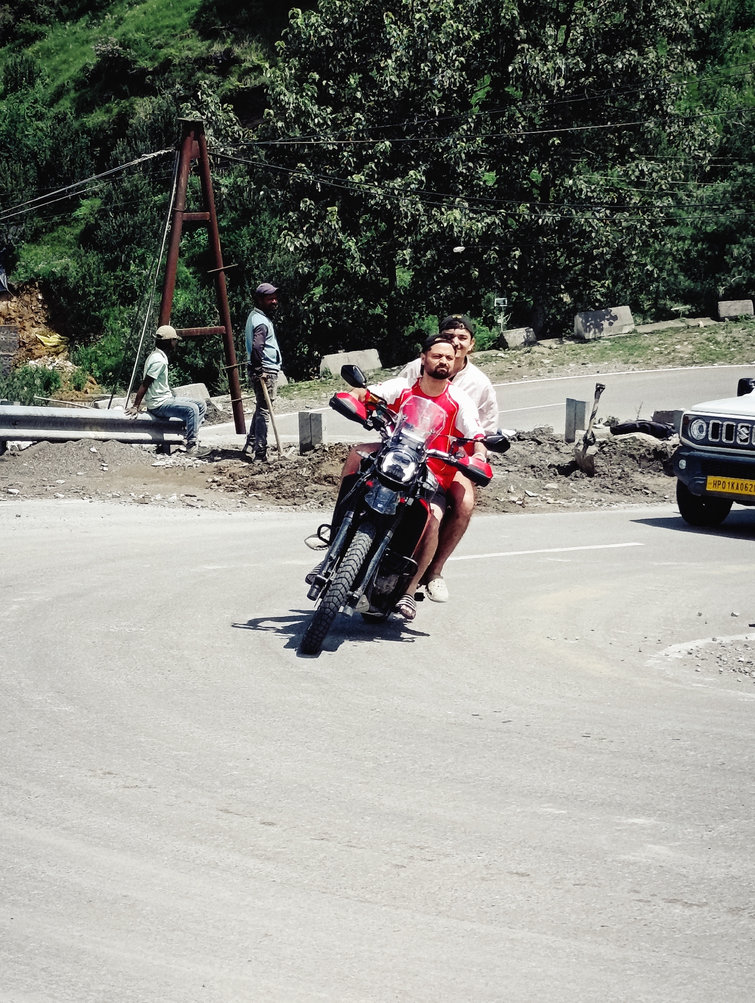 Two men are riding a red motorcycle on a paved road. The man in the front is wearing a red and white shirt and red shorts, while the man in the back is wearing a white shirt and a baseball cap. They are in Palchan, India. The road appears to be on a mountain slope, with a steep, vegetated hillside to the left and a dense cluster of trees behind the road. There is a metal guardrail on the left side of the road. In the background, a white SUV is parked on the side of the road, and construction materials are visible. Two other men are standing by the roadside near the guardrail, one of whom is holding a shovel. One man is wearing a blue shirt and a cap, and the other is wearing a light green shirt and a cap, and is sitting on the guardrail.