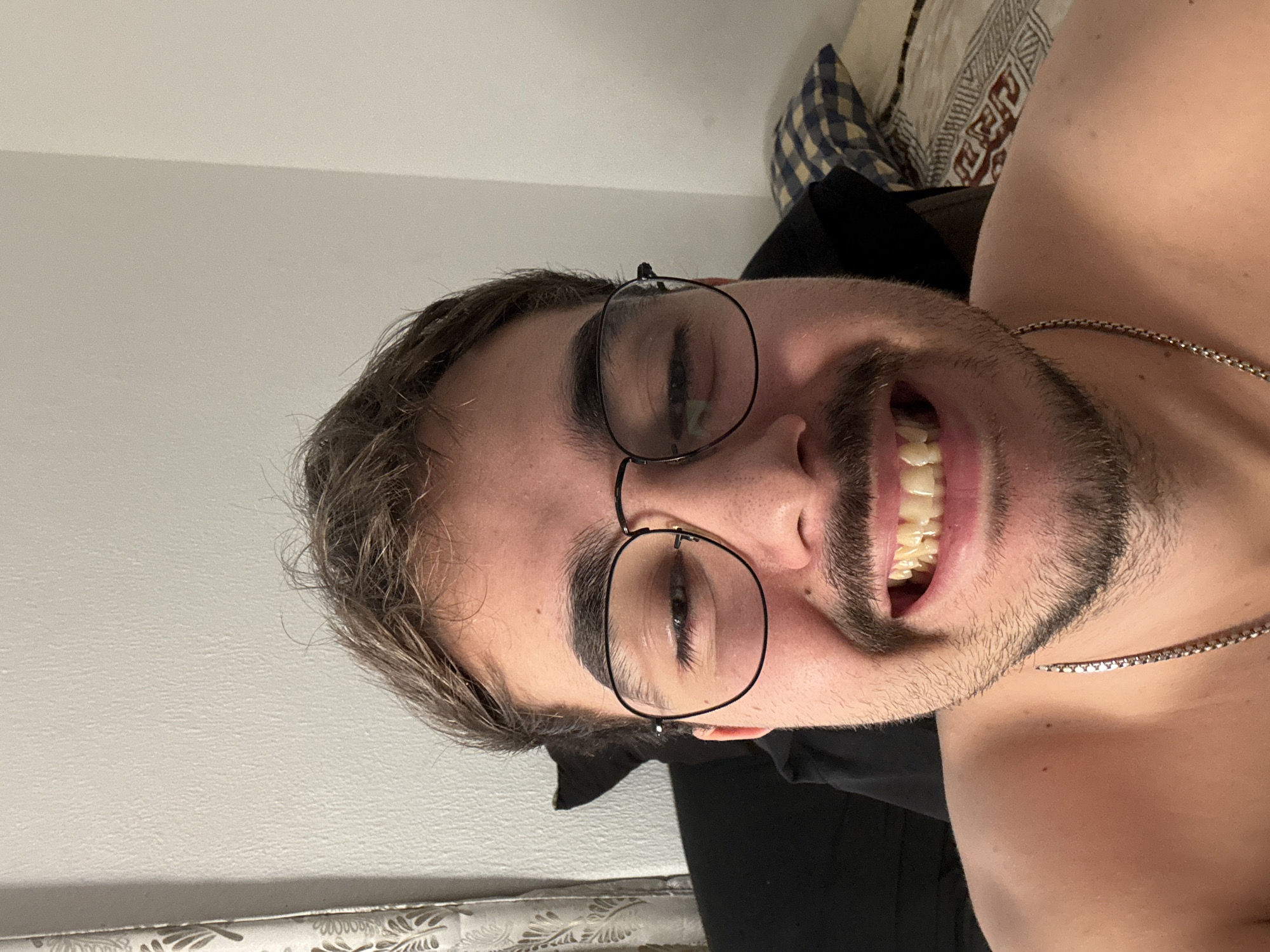 A close-up, chest-up selfie of a light-skinned man with dark, messy hair, a mustache, and a beard. He is wearing round, black-rimmed glasses and a silver chain necklace. He is smiling, revealing his teeth. The background consists of a white wall and a patterned blanket and pillow. The image was taken indoors, likely in a bedroom. There is no identifiable information about the location in Castelo Branco, Portugal.