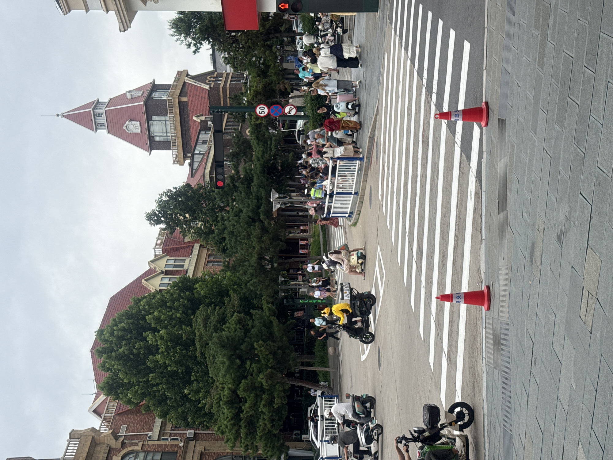 This image depicts a street scene in Qinhuangdao, China, likely on a cloudy day. The foreground features a pedestrian crosswalk with white stripes on asphalt. Two orange traffic cones, one with Chinese characters that appear to read "交通" (traffic/transportation), are placed on the sidewalk to the left and right of the crosswalk.
The middle ground is a bustling intersection with people and vehicles. Several individuals are riding motorbikes and electric scooters. One person in a yellow outfit rides a dark-colored scooter in the center. A group of people is gathered on a sidewalk to the right, some near a white railing. Traffic lights are visible, one showing a red light, and a circular speed limit sign indicates "50".
In the background, a distinctive building with a red, multi-tiered roof and a spire stands prominently. Adjacent to it are other brick buildings with red roofs, and several large, green trees line the street, providing shade. A white vehicle is parked on the left side of the street. The overall impression is of a busy urban environment with architectural influences from European styles.