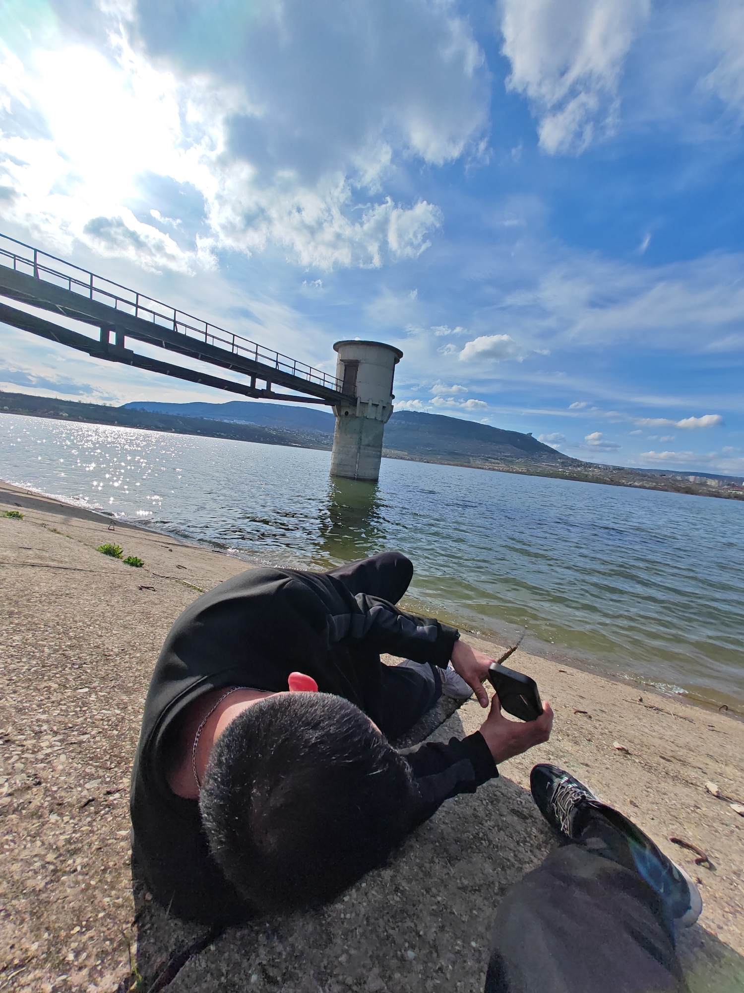 A medium, wide-angle shot captures a person lying on a concrete surface along a shoreline, with a body of water and a structure extending into the water in the background. The individual, dressed in dark clothing, is holding a mobile phone, and their head rests on the concrete. Visible is a pair of sneakers and pants.
In the background, a metal bridge or walkway leads to a cylindrical tower-like structure standing in the water. The water reflects sunlight, creating shimmering effects. Beyond the water, a hill or mountain is visible, and further in the distance, a cityscape is discernible. The sky is partly cloudy with patches of blue.
The location is the Lake Shumen in Bulgaria.