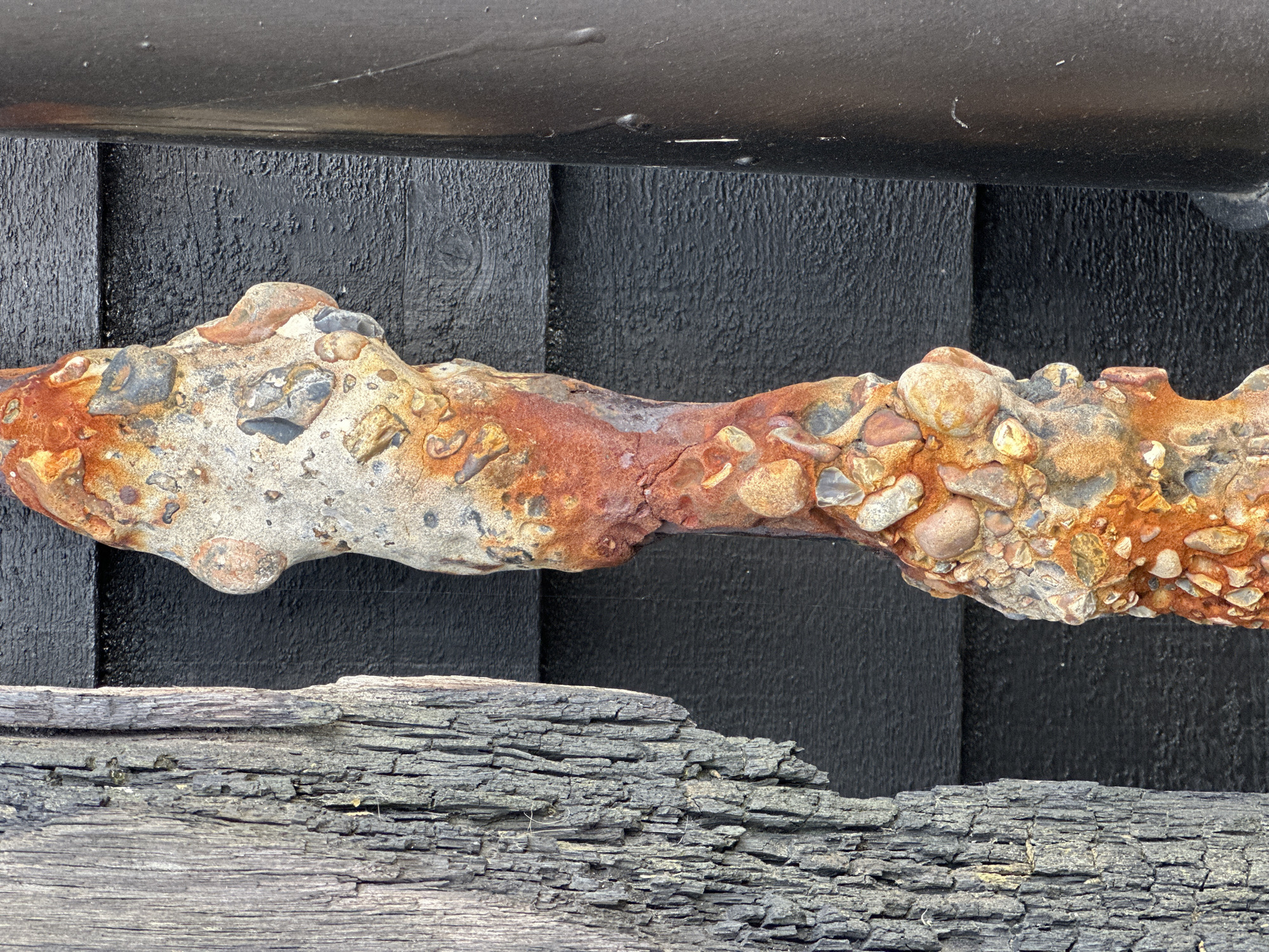 The image shows a close-up of a section of concrete and pebble-encrusted metal bar or pipe lying against a dark background composed of what appears to be painted wood. The concrete coating is heavily weathered, with exposed pebbles and rust-colored stains. Below the bar, there is a fragment of heavily weathered, possibly burnt, wood. The scene appears to be located outdoors. The precise location within Folkestone and Hythe District, United Kingdom, cannot be determined from the provided image alone.