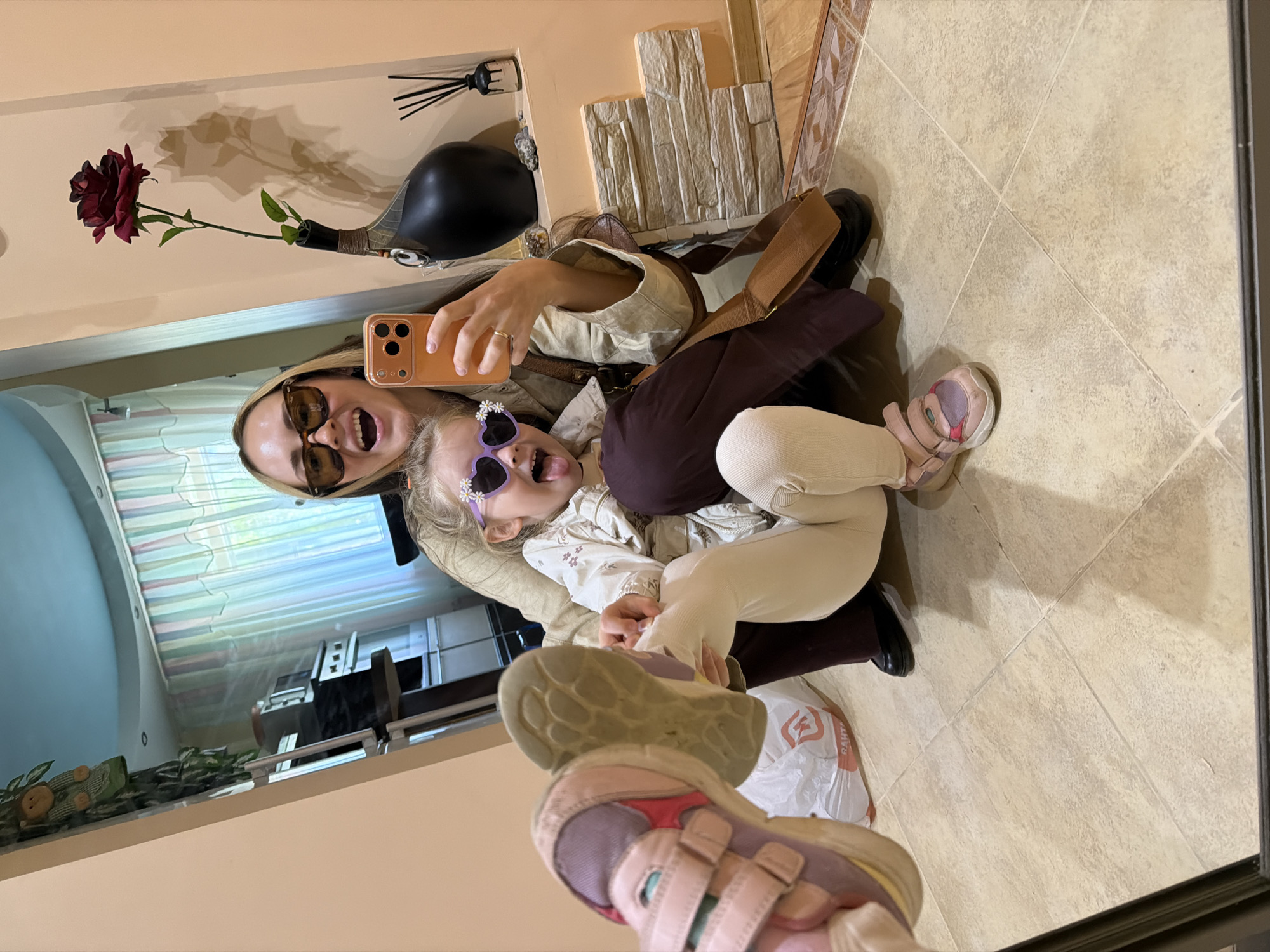 A woman and a child are taking a selfie in what appears to be a mirror. The woman has blonde hair, brown sunglasses, and is wearing a beige top. She is holding a mobile phone and smiling with her mouth open. The child, wearing sunglasses shaped like flowers, is sticking their tongue out. The background includes a wall with a vase containing a dark red rose, decorative stone bricks, and another vase with reeds. The floor is tiled in light beige. Reflected in the mirror is another room with what appears to be a window and various objects on a shelf. The location might be in Kamyshin, Russia, but there are no identifiable landmarks in the image to confirm this.