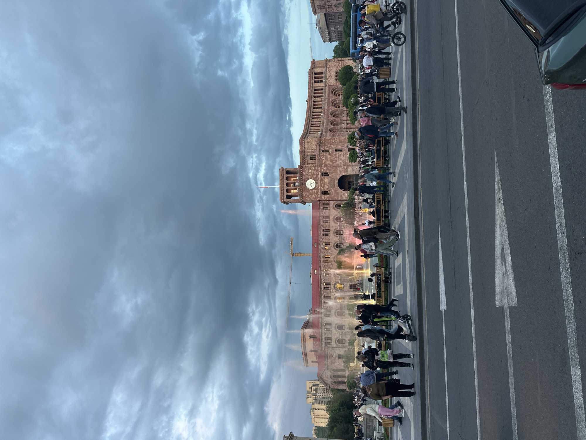 This image depicts a street scene in Yerevan, Armenia, likely in Republic Square, featuring the iconic government building with a clock tower. People are seated on benches along the sidewalk. A large crane is visible in the background near the building. The street has white directional markings. The sky is overcast with dark, heavy clouds. A dark-colored vehicle is partially visible on the right edge of the frame.