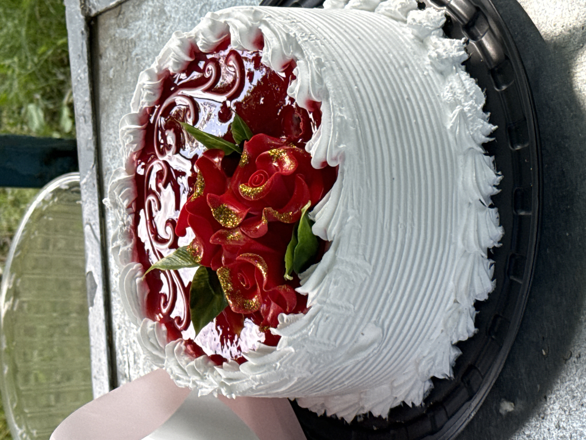 A round, white cake with red frosting roses and green leaves is displayed on a black plastic cake stand. The cake is decorated with white piped frosting around the edges and a swirl pattern on the top, topped with red glaze and decorative red roses. A pink ribbon is visible on the left side of the image. The cake is outdoors, placed on a metal surface with a background of greenery. No people are visible in the image. The location is described as Tsamakaberd, Armenia.