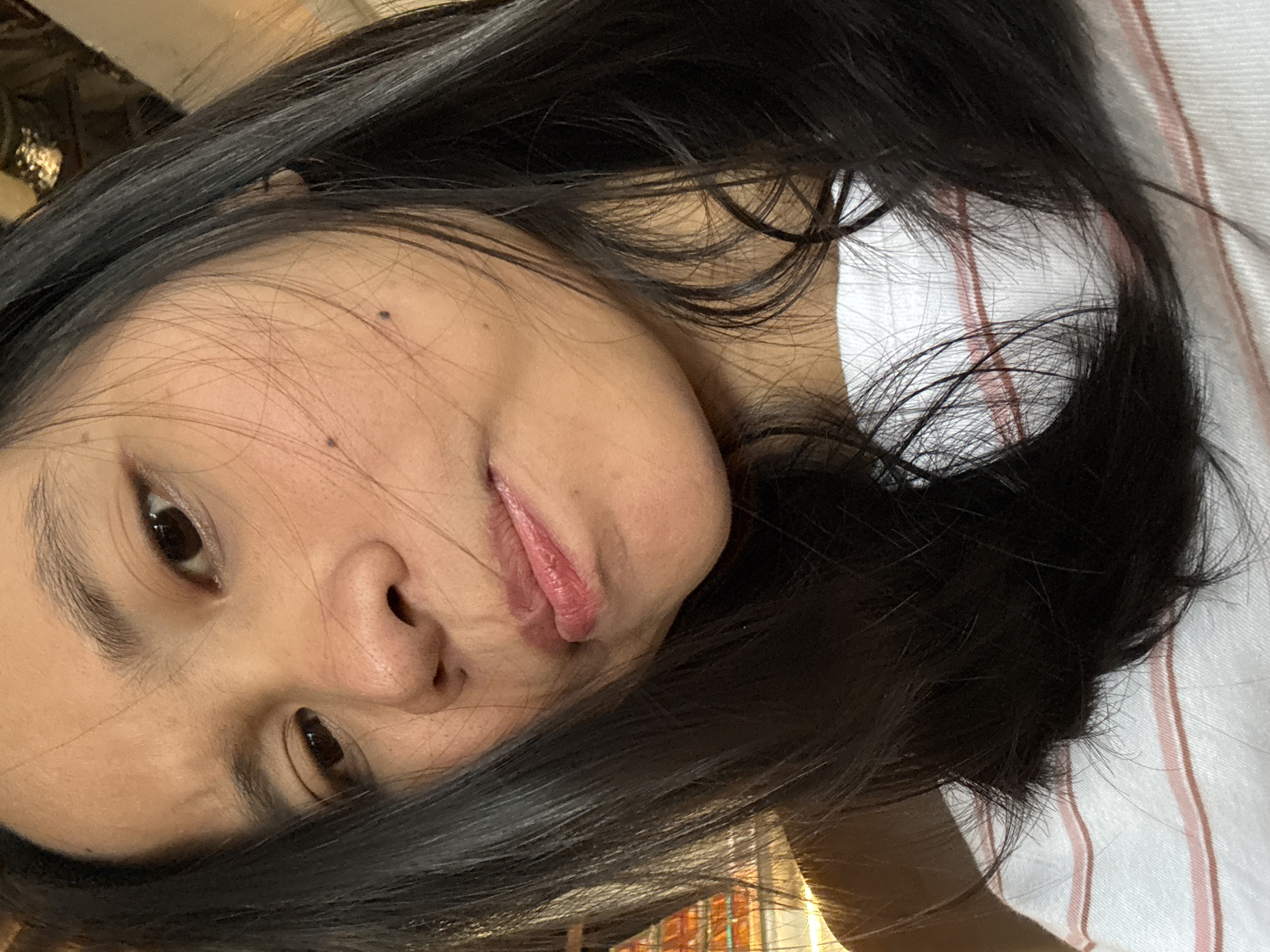 A close-up, low-angle selfie of a young woman with dark hair is depicted. She is looking towards the camera with a slightly pursed expression, her lips slightly parted. Her hair is dark and falls around her face, partially obscuring her shoulders. She is wearing a white t-shirt with thin pink stripes. The background is blurred, but appears to be indoors with some hints of light sources and textures. This image was taken in East Jakarta, Indonesia.