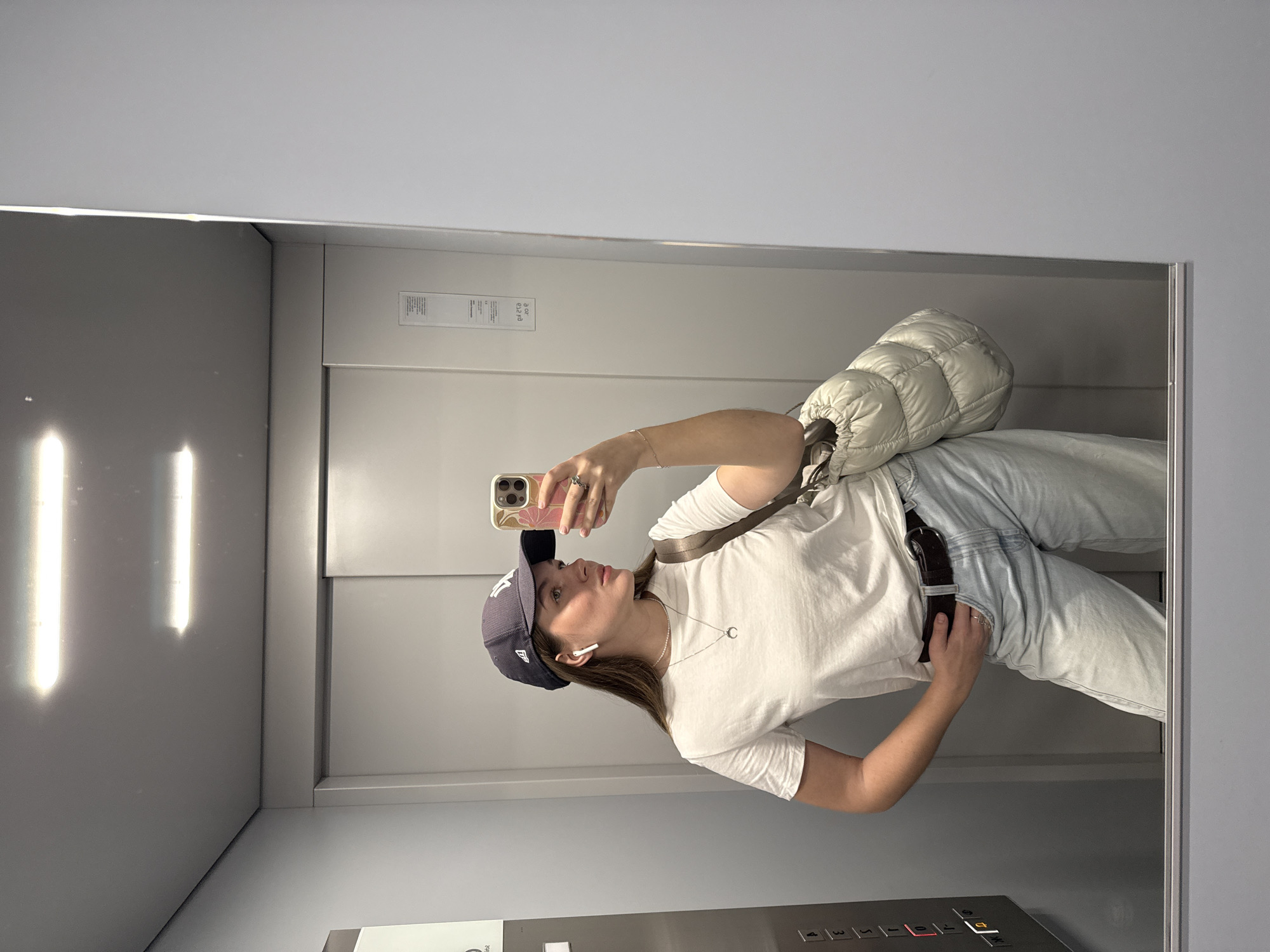 A woman is taking a selfie in an elevator. She is wearing a white T-shirt, light blue jeans, and a dark blue baseball cap. She has a white quilted bag slung over her shoulder and a brown belt around her waist. She is holding her phone up in her right hand and looking at the screen. The elevator is modern and clean, with gray walls and a metal control panel. The reflection in the mirror makes it appear that the image is rotated to its side. The location appears to be indoors and unlikely to be identifiable to Vozdovac Urban Municipality, Serbia, based on provided data.