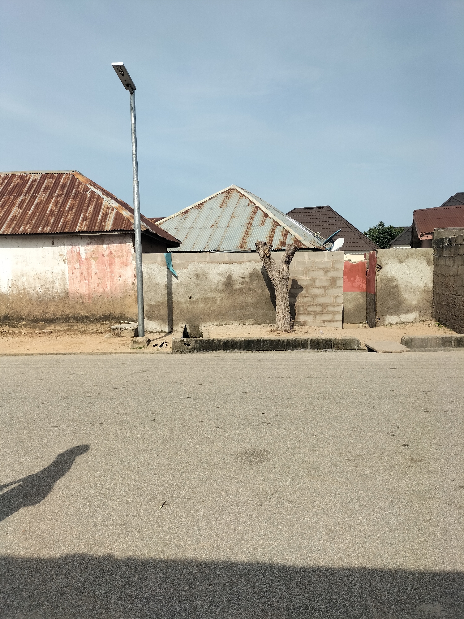 The image captures a street scene in Jalingo, Nigeria, featuring a paved road in the foreground with a distinct shadow of a person on the left side. Behind the road, a concrete wall, weathered and stained, serves as a backdrop. Several buildings with corrugated iron roofs, some showing signs of rust, are visible beyond the wall. A tall, modern street lamp stands prominently to the left of the wall. A bare, dried tree trunk stands in front of the wall, casting a shadow. A small satellite dish is mounted on one of the buildings. The sky is clear and blue. The scene appears to be an outdoor residential or commercial area on a sunny day.