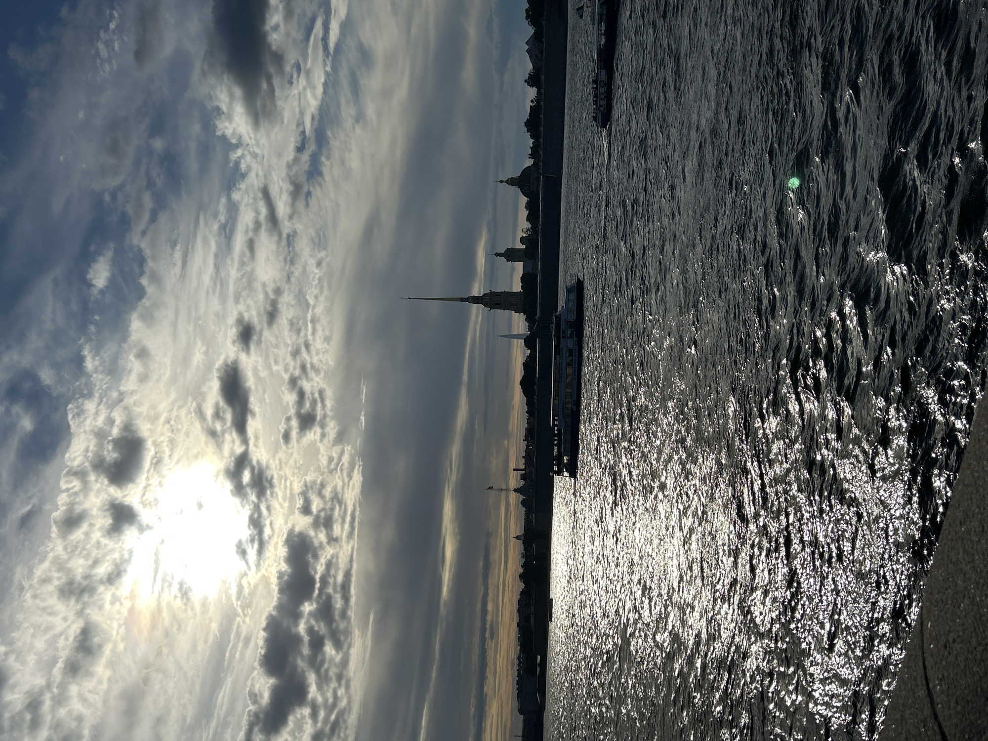 The image captures a wide view of a body of water, likely a river, under a dramatic sky filled with clouds. Sunlight pierces through the clouds, creating bright glares on the rippling water's surface. In the distance, a city skyline is visible, characterized by silhouetted buildings and a prominent spire. Several boats are present on the water: one larger vessel with multiple decks is moving from right to left, and a smaller boat is seen further in the distance on the left. A stone embankment is visible in the foreground on the left side of the image. The overall impression is one of a dynamic day with light and shadow playing across the scene.