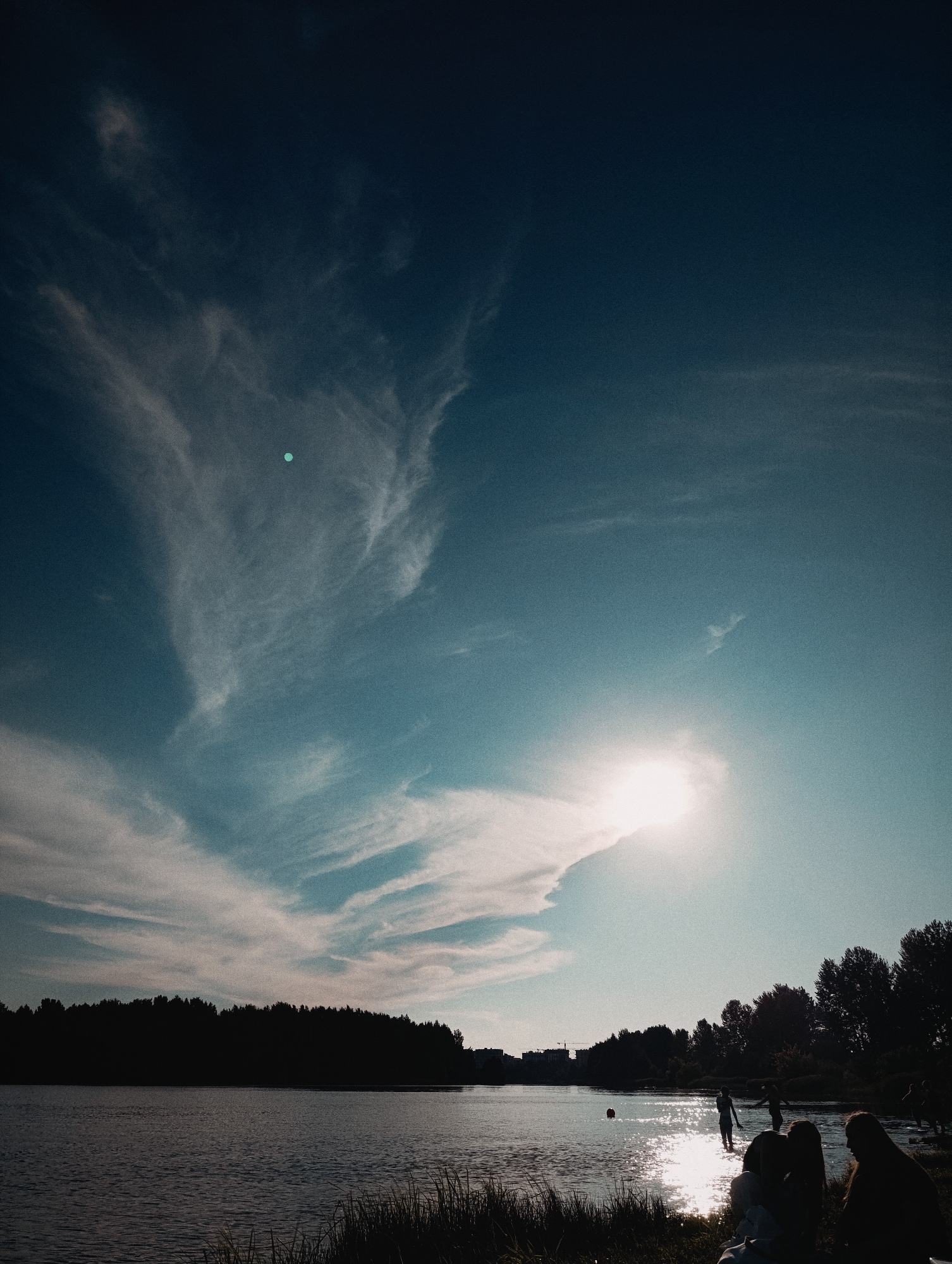 A wide shot captures a calm body of water reflecting the bright sky, with a dark silhouetted treeline on the far bank. The sky is a gradient of deep blue at the top, lightening to a bright cyan near the horizon, with wispy white clouds scattered throughout. The sun, a bright white orb, is partially obscured by a cloud in the upper right quadrant of the sky. In the foreground, tall grass lines the edge of the water. Several figures, silhouetted by the sun's glare, are scattered along the water's edge and in the shallow water. Some are wading, while others appear to be sitting or standing on the bank. A few distant buildings are visible on the horizon to the left of the treeline. The overall impression is of a peaceful, late afternoon scene by the water.