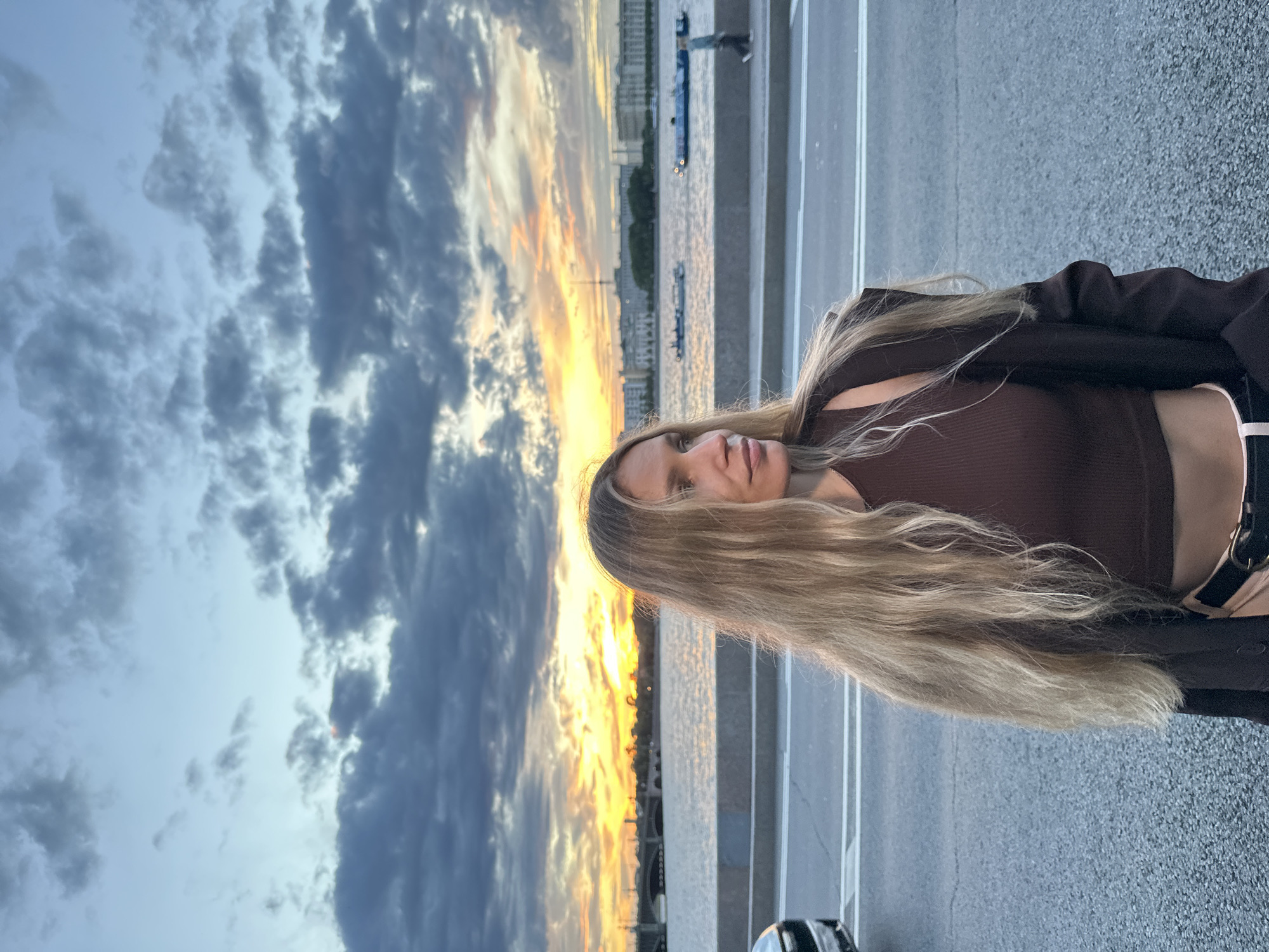 A young woman with long blonde hair is captured in a vertical, outdoor, medium shot. She is wearing a brown tank top and a dark jacket, with a bit of a pink waistband visible. Her gaze is directed upwards and slightly to the right.
The background features a dramatic sky filled with dark, textured clouds and vibrant streaks of orange and yellow from a sunset. Below the sky, a body of water, likely a river or canal, is visible with two small boats and what appears to be a city skyline with buildings and bridges in the distance. The foreground consists of a paved surface, possibly a road or promenade, with white lane markings. In the lower right corner, a car is partially visible.
The time of day is clearly sunset, with the sky ablaze with color. The weather appears to be transitioning, with dramatic cloud formations suggesting a dynamic sky. The presence of boats and the city skyline suggest an urban waterfront location, consistent with Saint Petersburg. The woman's upward gaze could imply contemplation or appreciation of the sunset. There is no visible text in the image.