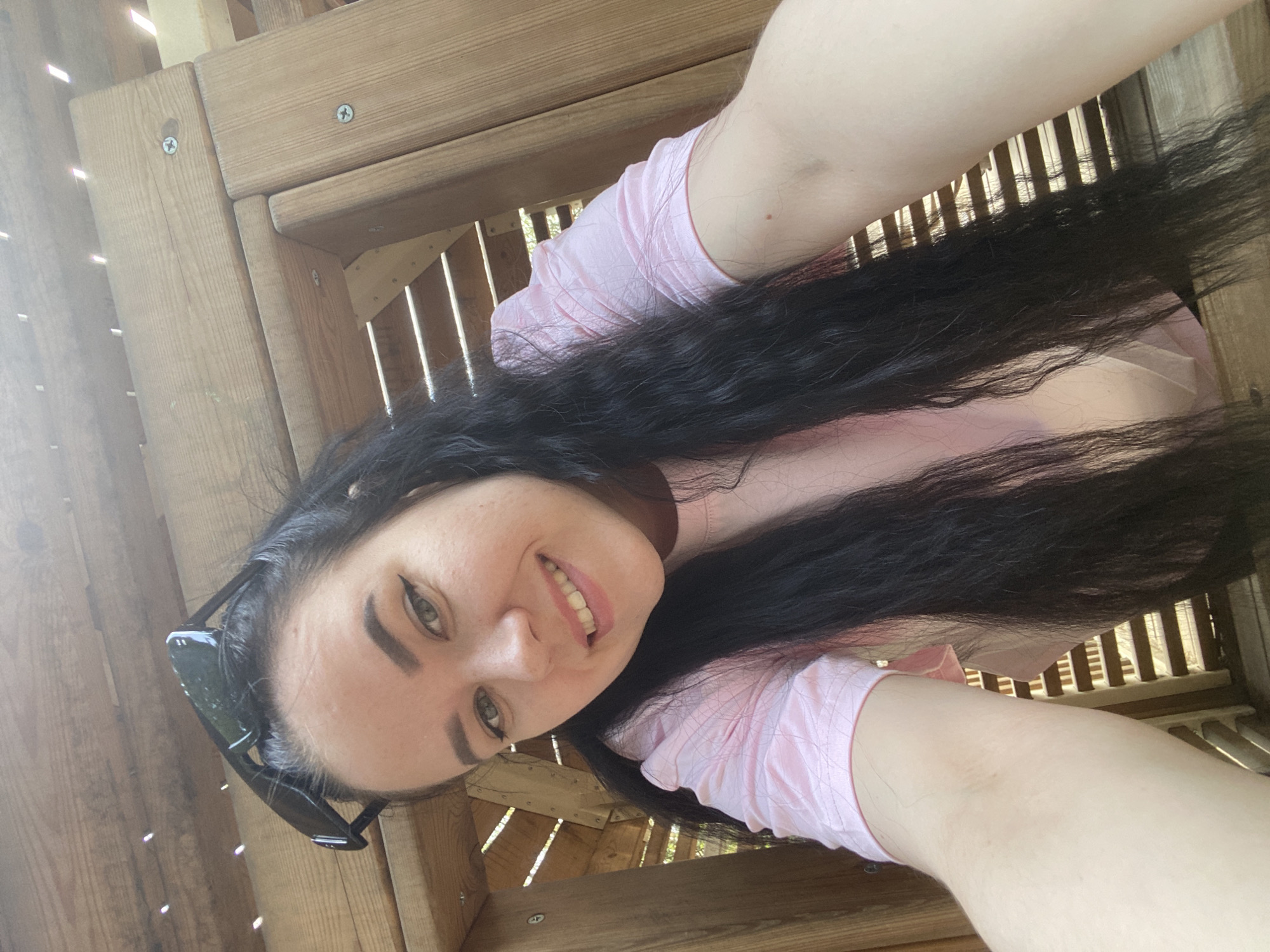 A selfie taken by a fair-skinned person with long, dark hair, wearing a pink shirt, is oriented at an angle against a wooden structure with slats. The individual is smiling, and sunglasses are visible on the left side of the frame. The setting appears to be outdoors, within a wooden play structure. There is no verifiable evidence to confirm that this image was taken in Perm, Russia.