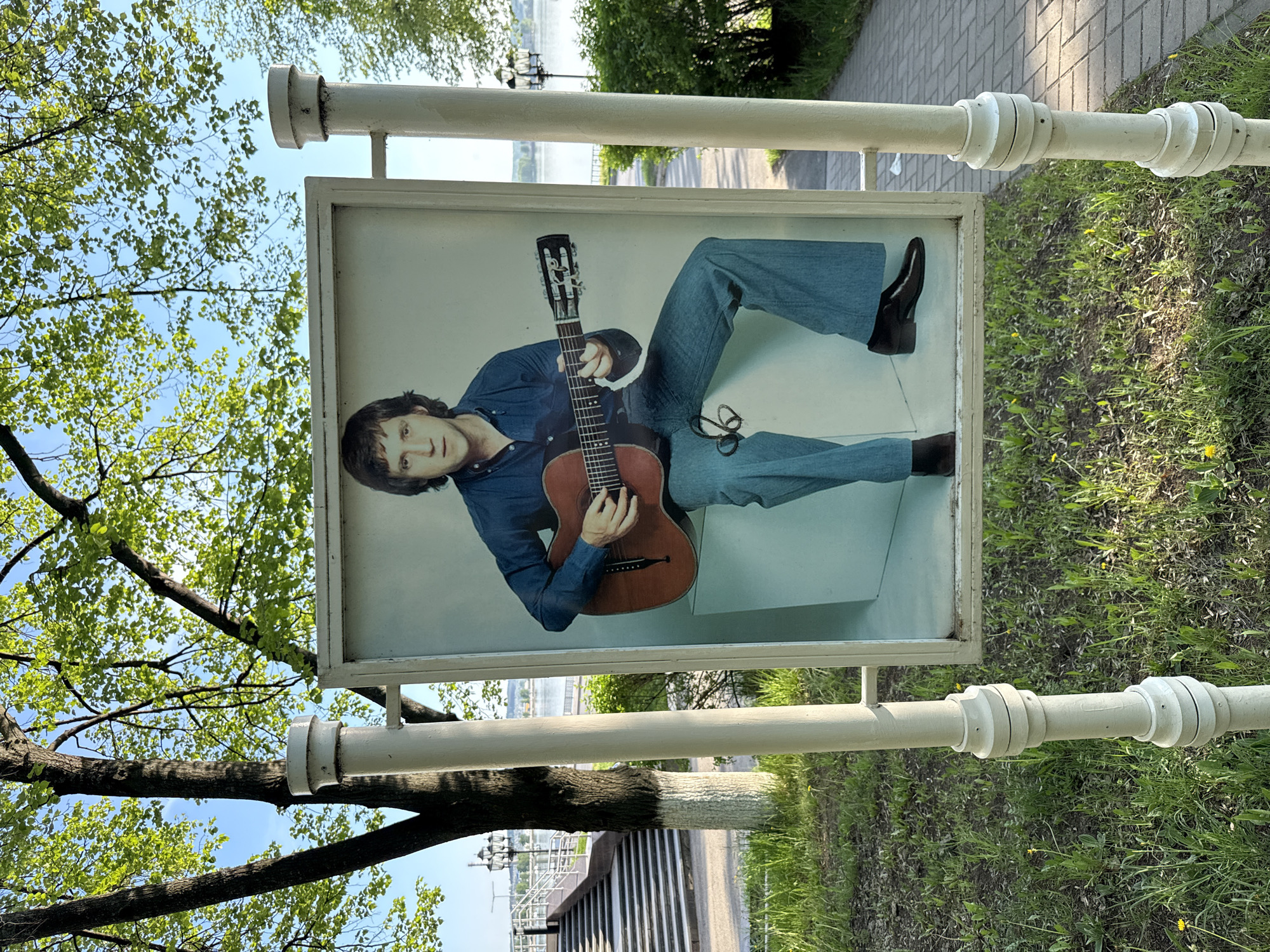 The image shows a poster of a man with long dark hair wearing a denim shirt and jeans, and playing a guitar. The poster is attached to a white metal fence, likely a memorial. The environment includes trees with green leaves and grass, and what appears to be a river or lake in the background. A paved walkway is visible on the right side of the frame. The location is Votkinsk, Russia, specifically a memorial to the musician Yuri Loza.