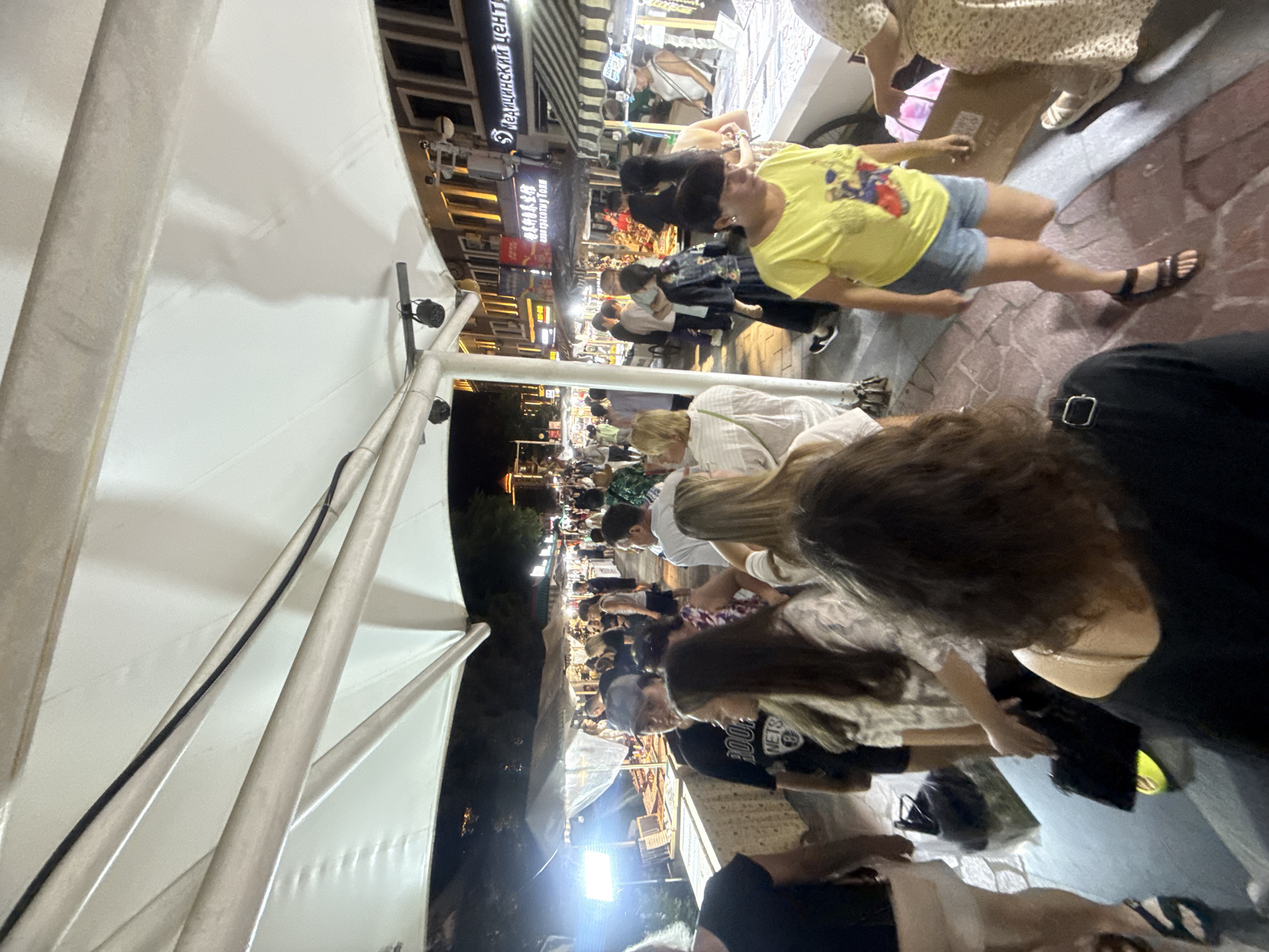 The image depicts a bustling night market in Qinhuangdao, China, likely at a street stall. A white awning stretches overhead, providing shade and support for lights and wires. In the foreground, several people are visible, some walking and others browsing stalls. A woman in a yellow t-shirt and denim shorts is prominent, looking towards the right. Another person in a black shirt is in the lower right corner, with their back to the camera. The market appears to have numerous stalls displaying various goods, with a crowd of shoppers navigating the narrow walkway. The overall atmosphere suggests a lively and busy commercial area at night.
