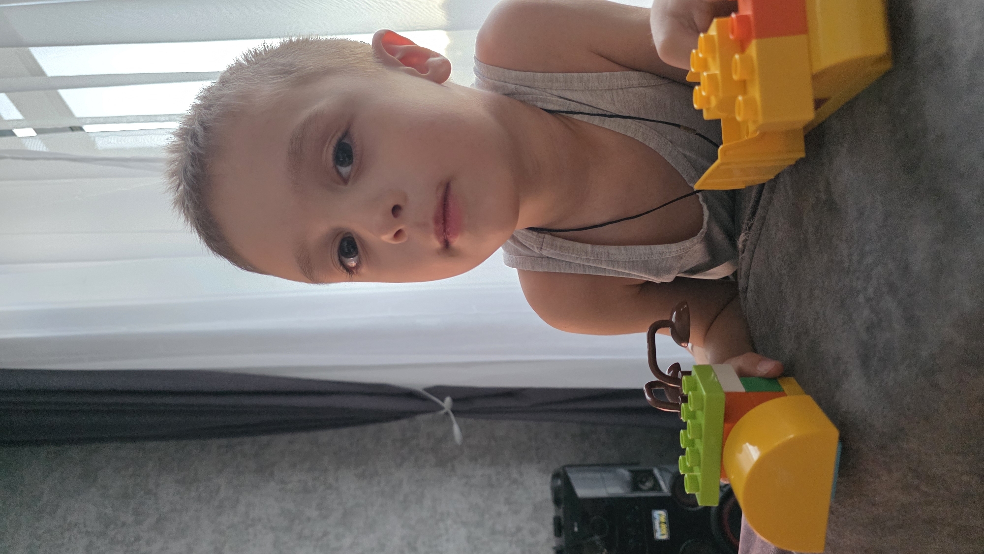 A child is lying down on a grey surface, likely a sofa or bed, playing with building blocks. The blocks are primarily yellow, orange, and green. The child is wearing a grey tank top. The background includes a window with white blinds and a dark curtain. A black object, possibly a speaker or electronic device, is also visible. The image does not contain information to confirm a location in the city of Bulgakovo, Russia.