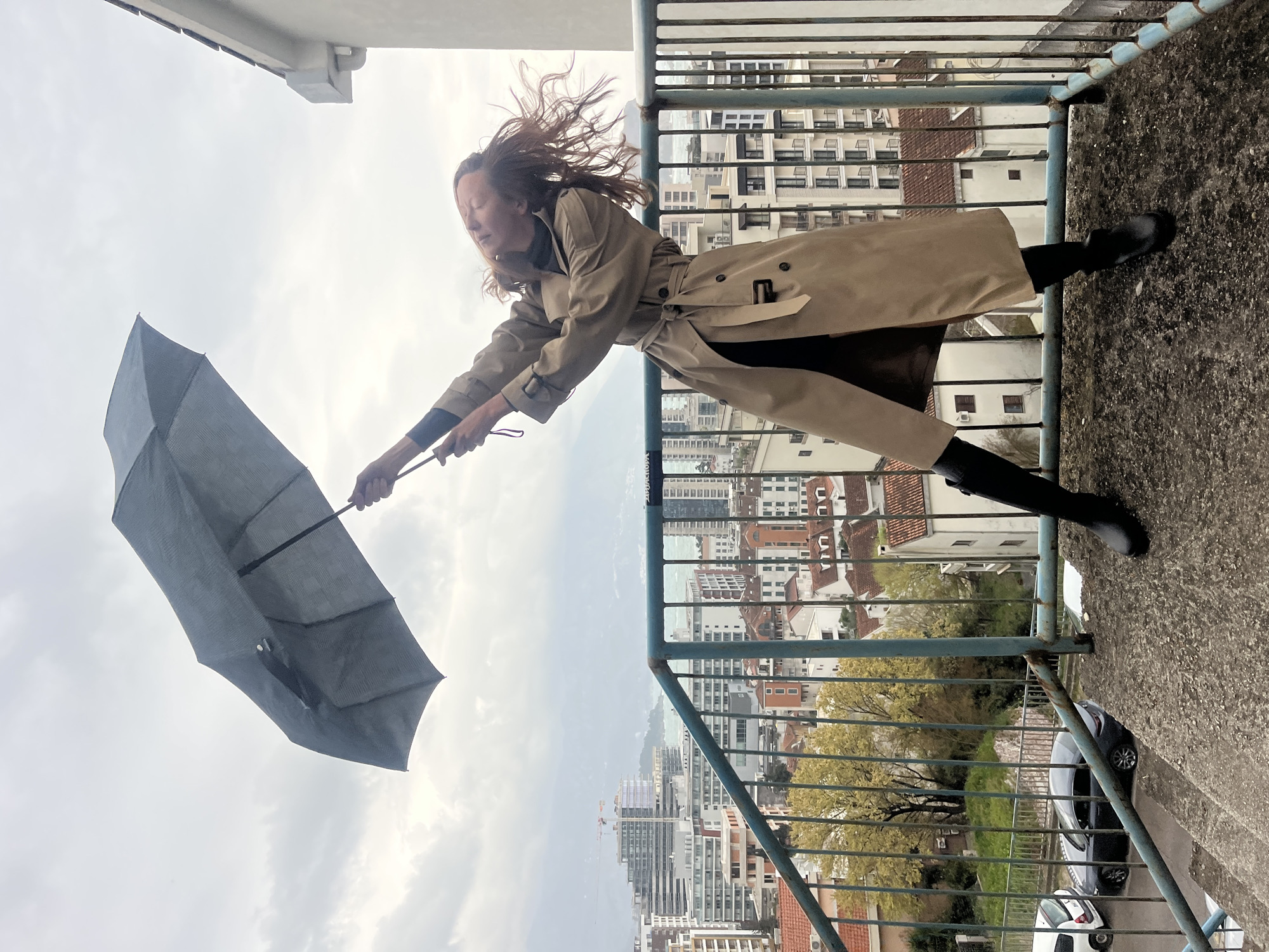 An outdoor scene depicts a female individual on a concrete landing overlooking the city of Budva, Montenegro, under an overcast sky. The individual, positioned slightly right of center, faces left, leaning backward and right, bracing against strong wind. Her long, light brown hair is disheveled and blown upwards by the wind, partially obscuring her face. She is dressed in a light brown trench coat, belted at the waist, and dark boots. With both hands, she grips a dark grey umbrella, which is forcefully pushed upwards and appears partially inverted by the wind. A weathered, blue-painted metal railing, showing signs of rust, runs horizontally in the mid-ground, separating the individual from the panoramic city vista.
Below, the urban landscape of Budva extends, featuring a dense mix of modern multi-story buildings, some under construction with visible cranes, alongside older structures characterized by red-tiled roofs. The Adriatic Sea, appearing choppy, is visible in the middle distance, with hazy mountains discernible on the horizon. Sparse trees with yellow-green foliage and a road with parked vehicles, including a white car and a dark sedan, are visible at a lower elevation within the city. A portion of a light-colored building with a dark roofline and white gutter is visible on the upper right.
