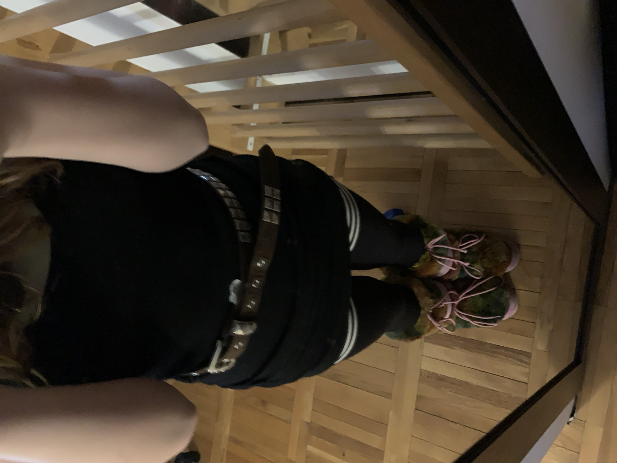 The image shows a person lying underneath a wooden slatted structure, possibly a bed. The person is wearing black clothing, leggings with white stripes, a studded belt, and furry sneakers with pink laces. The flooring appears to be wooden planks. The location cannot be determined from the image.
