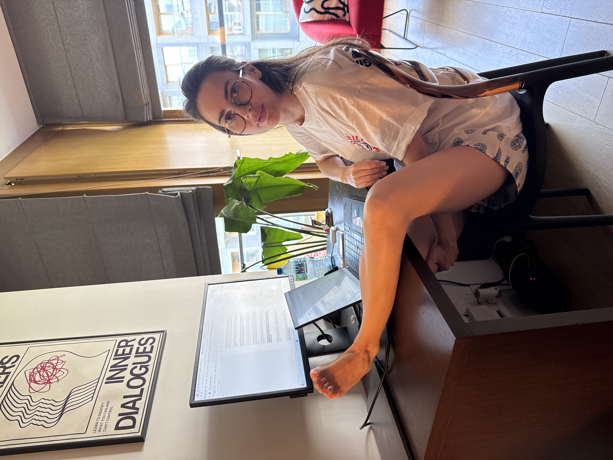 A woman is sitting in a black chair at a dark wooden desk. She is wearing round glasses, a white t-shirt, and patterned shorts. Her right leg is resting on the edge of the desk, near a computer monitor displaying text. There is a smaller screen propped up behind the main monitor, along with a water bottle and a plant. A poster with the words "INNER DIALOGUES" is on the wall to the left of the monitor. The background shows a building through a window. The location is in the city of Unknown, Unknown.