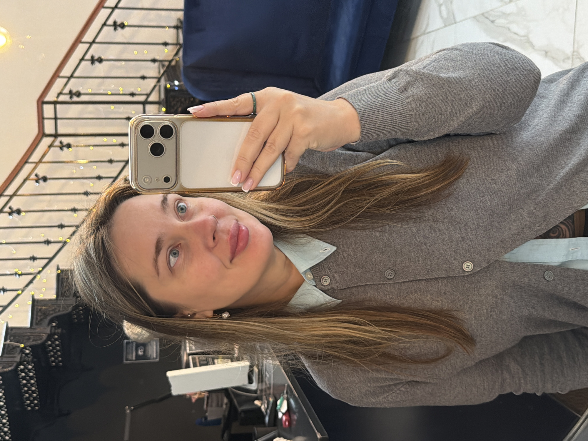 A fair-skinned woman is taking a selfie with a gold-cased smartphone. She has blue eyes and light brown hair with gray roots. She is wearing a gray cardigan over a light blue collared shirt and an emerald green ring on her right hand. The background includes a staircase with black railings and a blue cushion. The picture also appears to show part of a retail display case, suggesting the woman may be in a store. Based on the ornate style of the staircase and the presence of a jewelry display, it is plausible that the location could be inside a high-end department store or luxury retail space in Moscow, Russia, given the presence of such establishments there.