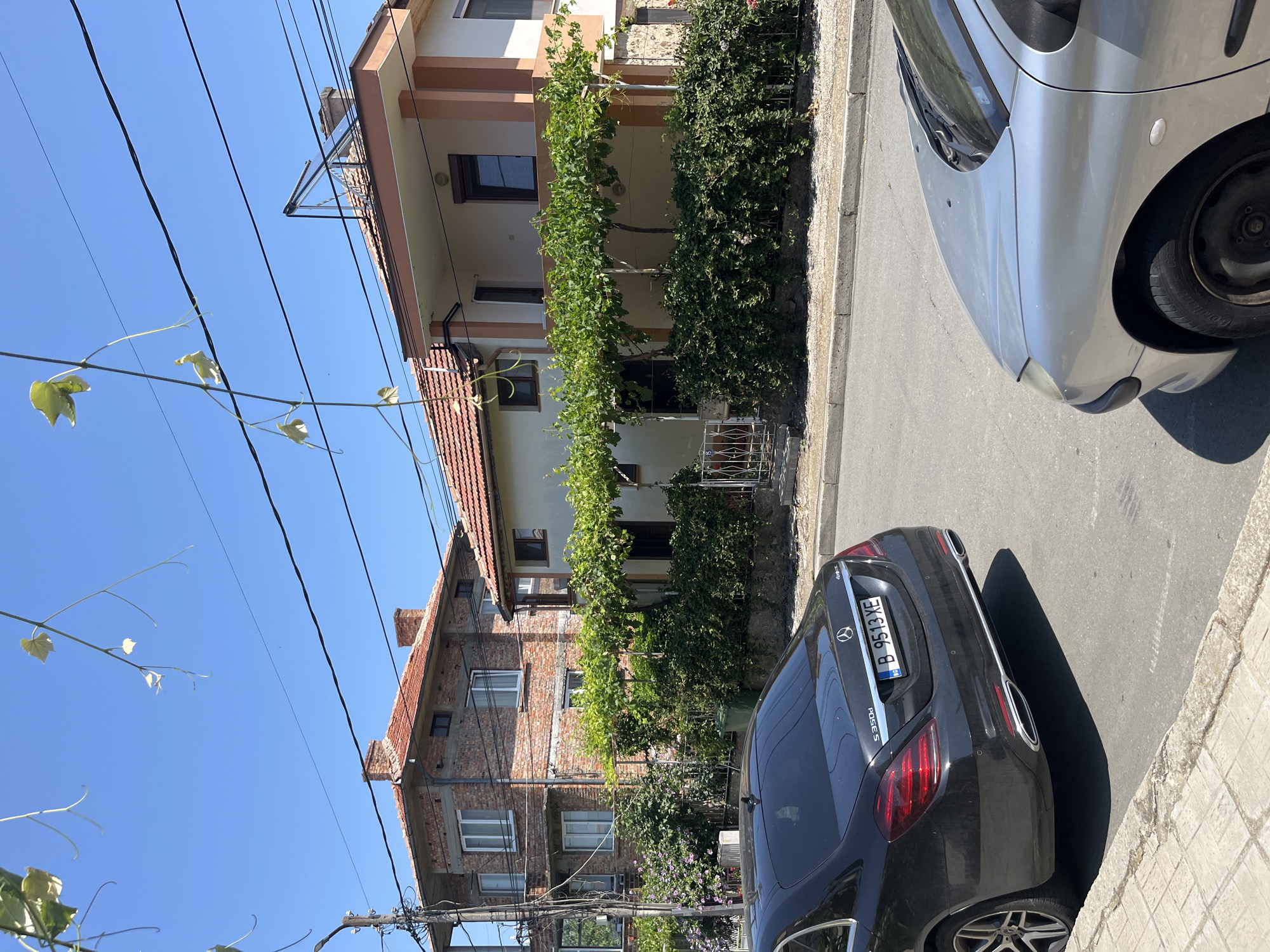 The image depicts a street scene in Obzor, Bulgaria, on a clear, sunny day. In the foreground, a black Mercedes-Benz S350d is parked on the right side of the road, with its rear end facing the camera. To the right of the Mercedes, the front fender and hood of a silver Peugeot are visible. The street is paved with asphalt, and a concrete sidewalk runs along the left edge of the road.
Behind the parked cars, two houses are visible. The house on the left is a two-story brick building with a red-tiled roof and white-framed windows. The house on the right is a two-story structure with a lighter colored facade, also featuring a red-tiled roof. Both houses have overhanging eaves supported by posts, with lush green vines growing over them, creating a shaded area. A small white metal gate is visible in front of the right house.
The environment is characterized by residential buildings, vegetation, and infrastructure typical of a European town. Power lines crisscross the sky above the houses. The overall impression is a quiet, ordinary street on a summer day. There are no people visible in the image.