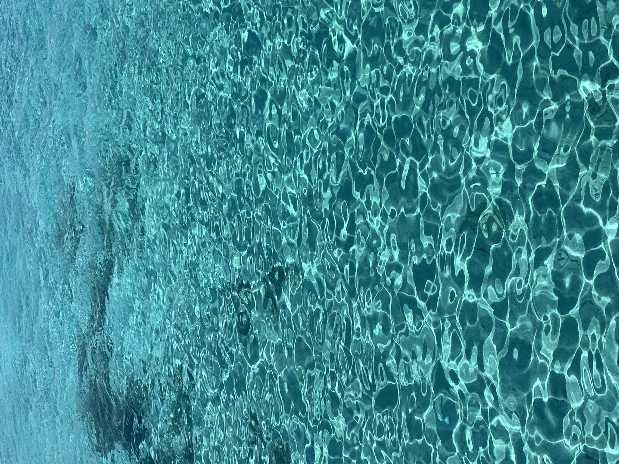 The image displays a close-up, overhead view of clear, turquoise water with visible ripples and light refractions. The water appears shallow, and the seabed is partially visible in darker, shadowy areas towards the top left of the frame. There are no people, subjects, or objects in the image, nor any discernible actions or interactions. The environment is solely the surface of the water, with no indication of a specific urban or natural setting beyond what can be inferred from the water's clarity and color, which is characteristic of coastal or tropical locations. The location in Neo Chorio, Cyprus, is not visually represented in the image itself.