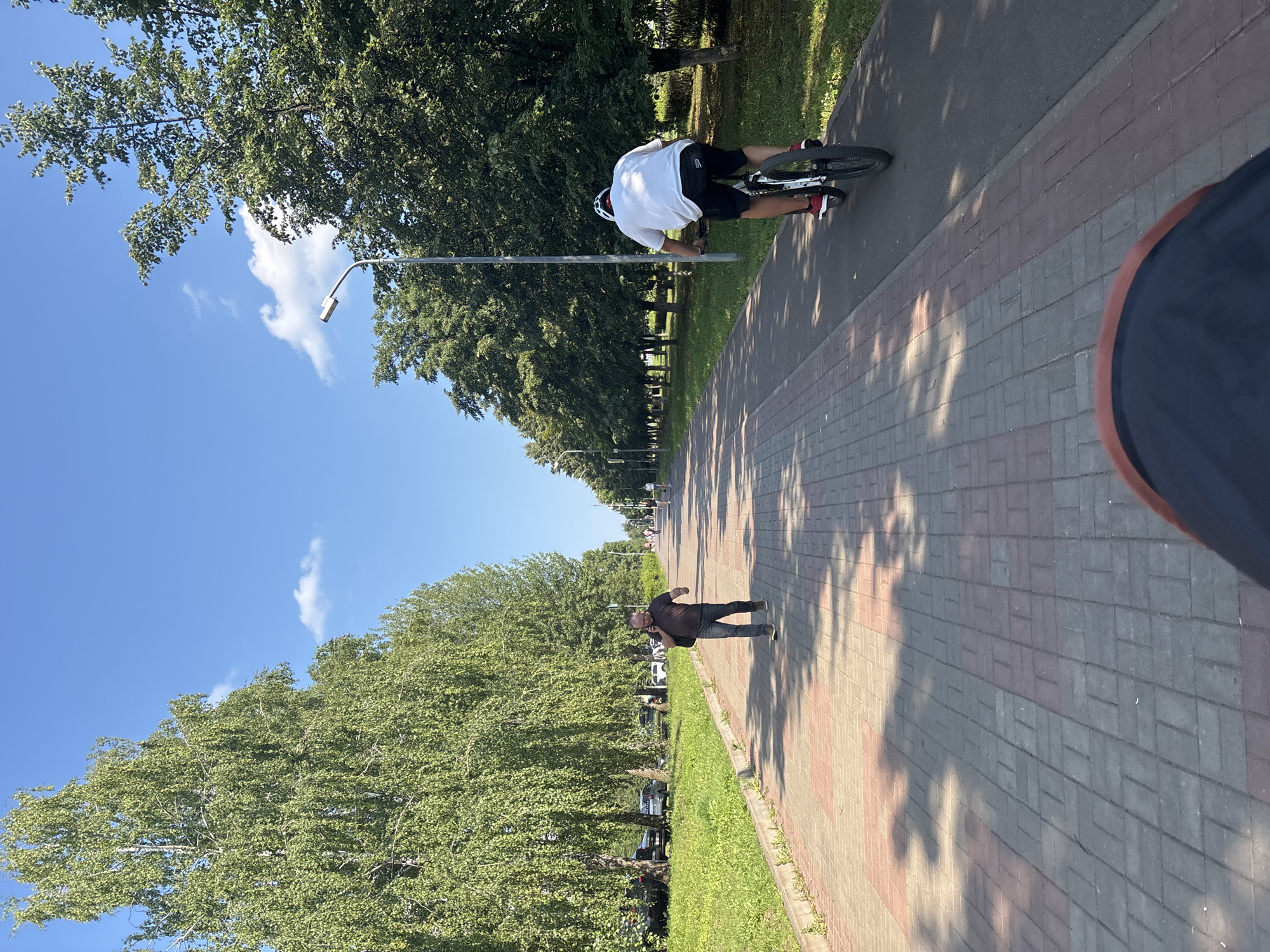 This image depicts a sunny day in a park-like area with a paved walkway. The walkway is made of interlocking bricks in a pattern of red and grey. On the left side of the walkway, there is a grassy area with several cars parked along a road, bordered by tall, leafy green trees. On the right side of the walkway, there is a paved cycling path.
In the foreground, a person's legs and feet are visible, suggesting the photo is taken from someone sitting or kneeling. Further down the walkway, a man in a dark t-shirt and jeans is walking and talking on a cell phone, carrying a dark bag over his shoulder. He appears to be walking towards the viewer. Beyond him, several other people are walking or cycling in the distance.
A cyclist in a white t-shirt and black shorts, wearing a white helmet, is riding a bicycle on the cycling path towards the viewer. The cyclist has red shoes.
Tall, mature green trees line both sides of the walkway and cycling path, casting dappled shadows on the ground. The sky is bright blue with a few scattered white clouds. A street light is visible on the right side of the pathway, near the cyclist.
Based on visual cues such as the architecture of the streetlights, the style of the trees, and the general urban park environment, the location is identified as Naberezhnye Chelny, Russia. This specific area appears to be a well-maintained public space designed for recreation and pedestrian/cyclist traffic.