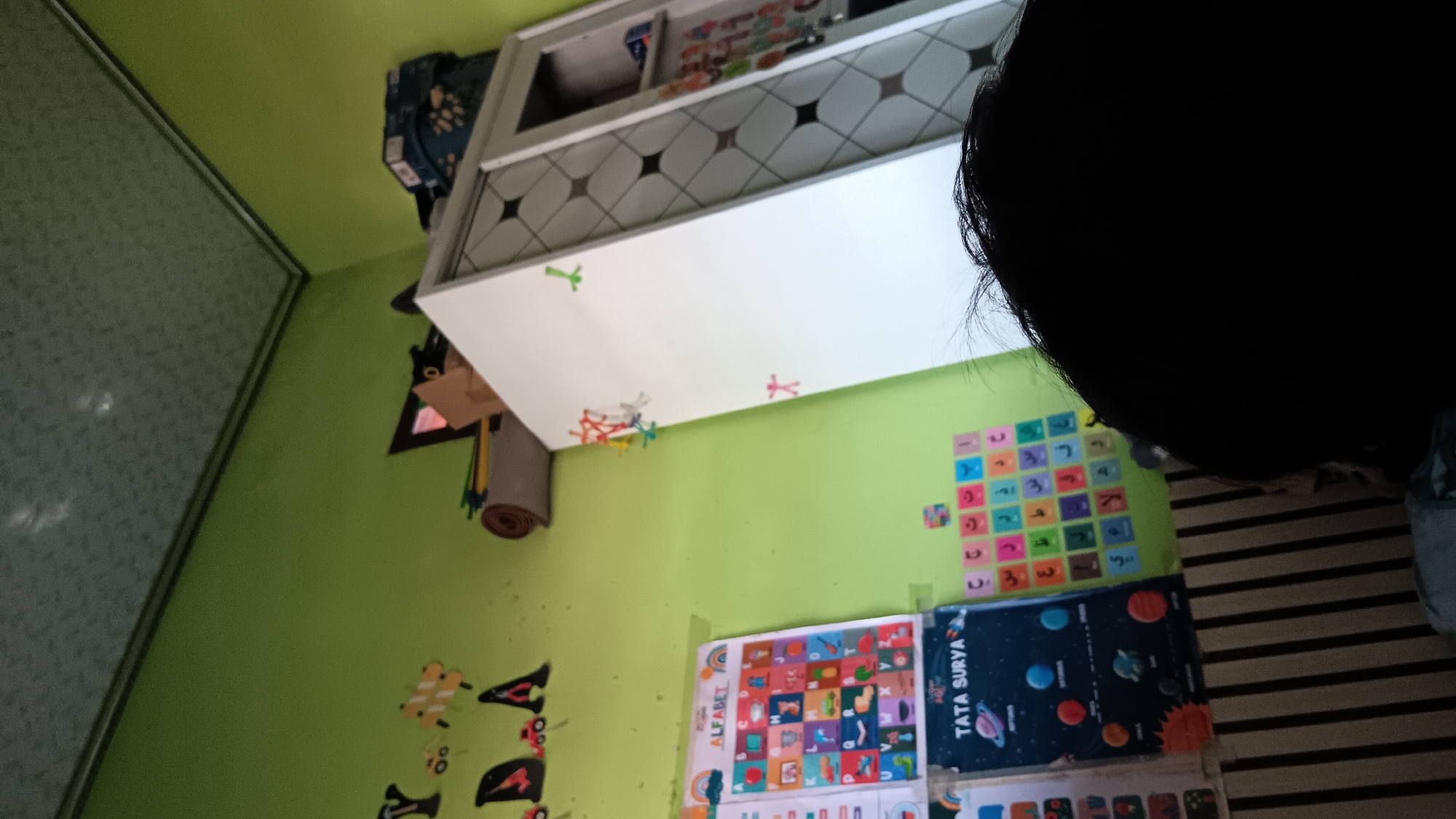 The image depicts a child's room, likely in Sonobekel, Indonesia, with green walls and a textured ceiling. A white wardrobe stands against the right wall, adorned with colorful stick figures. To the left, a chart labeled "ALFABET" with various images and letters is taped to the wall, alongside another chart showcasing planets titled "TATA SURYA." Below these, a chart with Arabic letters is visible. Various toys and decorations, including a rolled-up mat and colorful cutouts of fish and letters, are scattered around the room. In the foreground, the back of a child's dark hair obscures a portion of the scene.