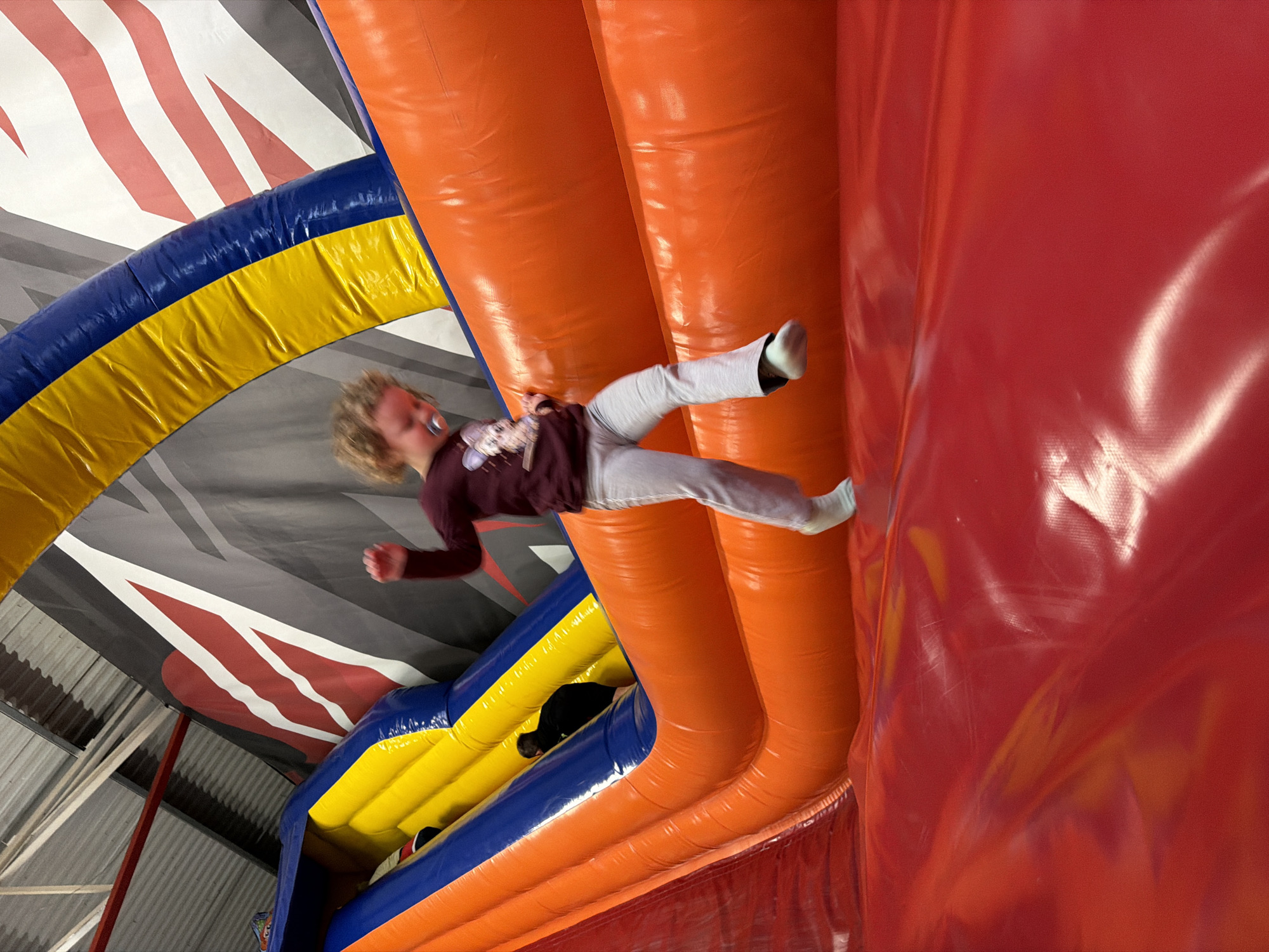 The image shows a child appearing to be suspended in mid-air within an indoor inflatable play structure. The child is wearing a maroon long-sleeve shirt, light gray pants, and white socks, and has fair skin and light hair. A pacifier is visible in their mouth. They appear to be in motion, with one fist clenched.
The play structure is made of brightly colored inflatable components: blue, yellow, orange, and red. Sections of the structure are inflated into cylindrical shapes. The background shows the ceiling of the building, which has a gray color with red and white markings.
The image was taken in Merzig, Germany.
