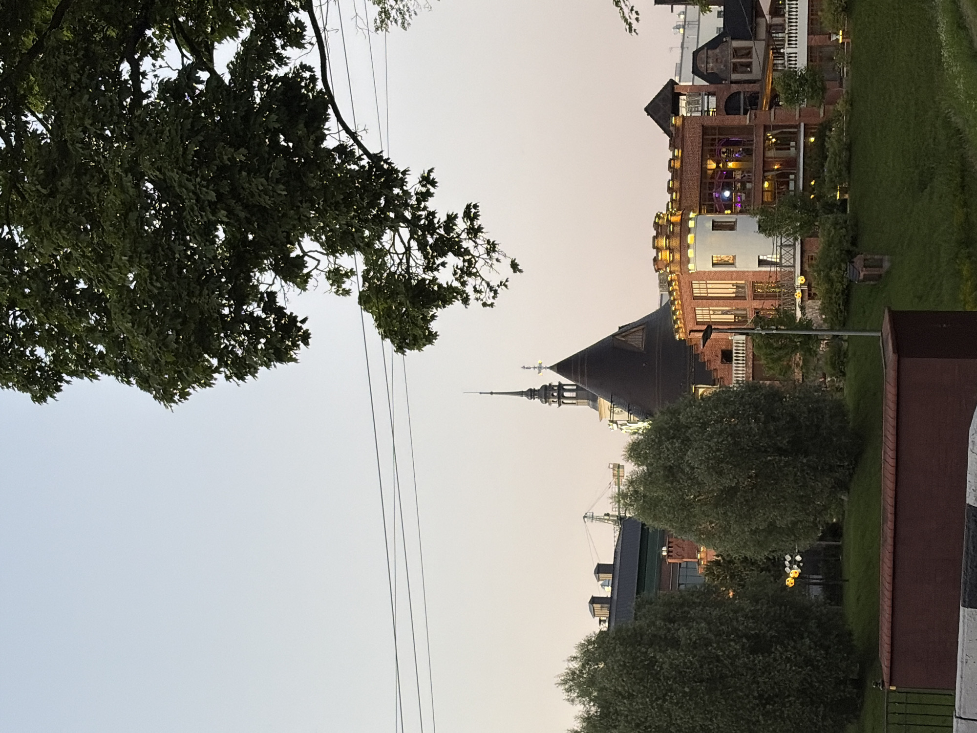In the city of Unknown, Unknown, a large tree branch with green leaves hangs from the top right corner of the frame, obscuring the pale sky and some electrical wires stretched across it. Below the sky, a dark, conical roof structure is visible, topped with a slender spire that has a cross. To the right of this, a multi-story building with large windows and decorative lighting is illuminated from within, casting a warm glow. This building is constructed of red brick and features crenellated battlements and white railings. In the foreground, two large, dense trees with rounded tops stand on a grassy slope. A low brick wall runs horizontally across the bottom of the image, partially obscuring a black and white striped barrier.
