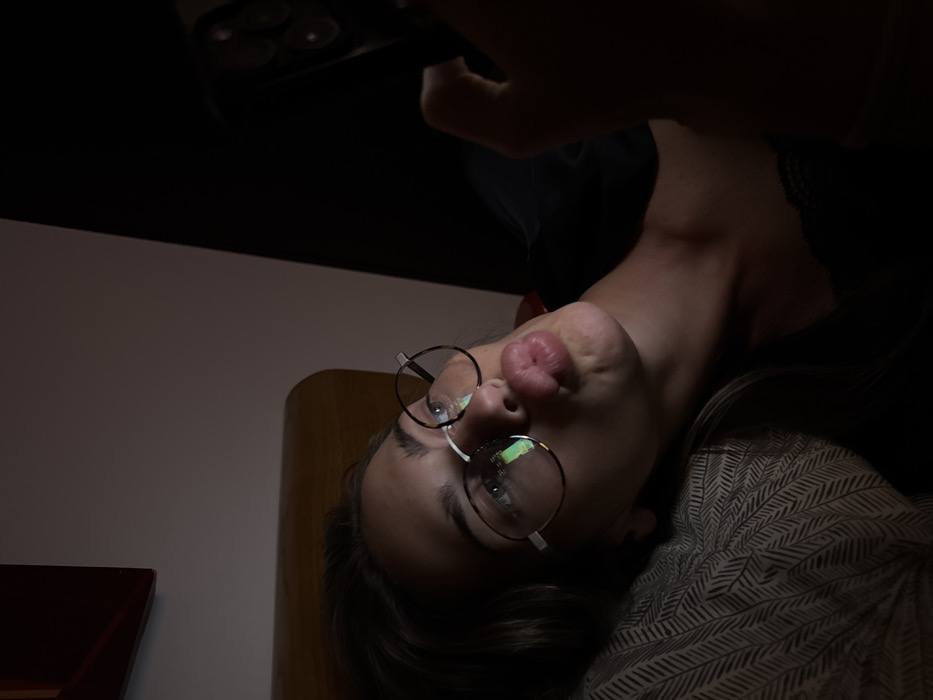 A woman is tilted upside-down, looking up. She's wearing glasses with round frames and has her lips pursed. Her dark hair blends with a dark patterned material. The background features a mix of light and dark surfaces. A hand holds a device, perhaps a phone, above her, reflecting light. The overall lighting is dim, casting shadows and highlighting the woman's face.