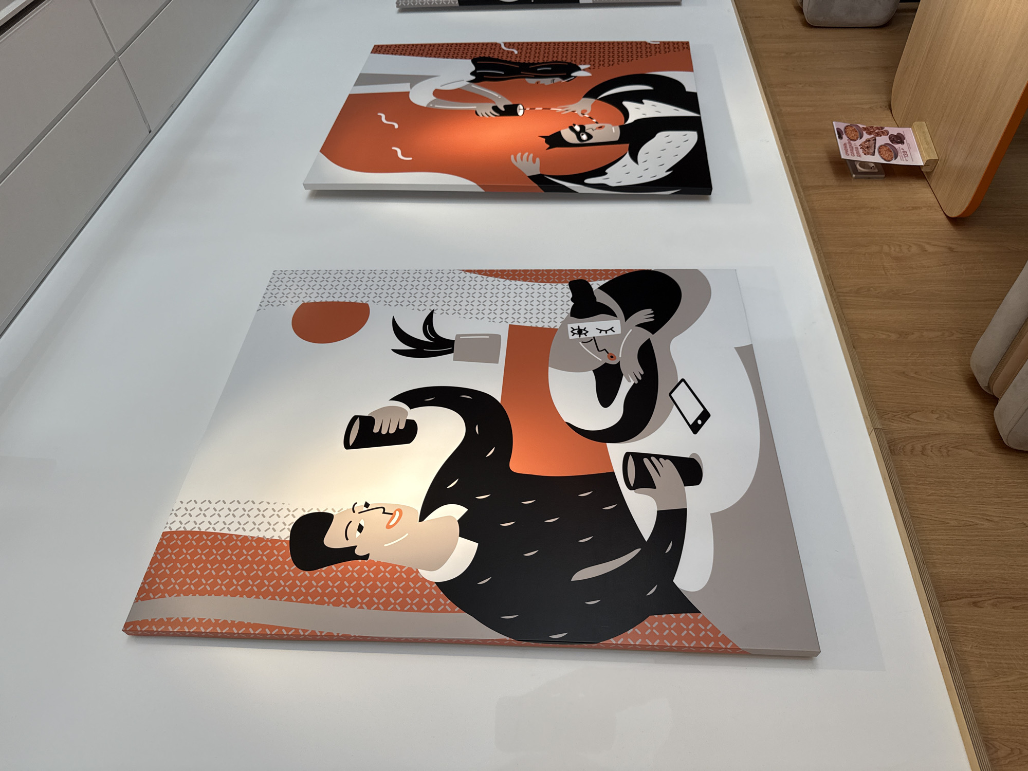 The scene shows two illustrative artworks displayed on a white wall. The top artwork depicts two figures in a stylized, contrasting color scheme of white, black, and orange. One figure, possibly a woman, holds a device resembling a flashlight or a phone, while the other figure is depicted with masked eyes and a cape-like shape, suggesting a superhero or a masked character. The orange background creates a sense of light or energy emanating from the device.
The lower artwork presents a similar stylized design with a man lying down, holding a device, while another figure sits nearby with eyes closed or masked. A smartphone is visible near the seated figure. The color palette is similar to the upper artwork, featuring white, black, orange, and grey.
A small price tag or advertisement is visible on a light-colored wooden floor near the right edge of the image. The context of the artwork and the presence of the advertisement suggests the scene is likely within a retail or promotional space. I have no information about Tobolsk, Russia.