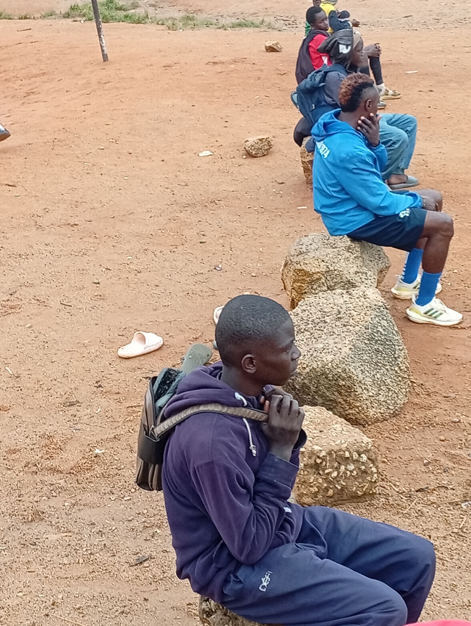 A group of young men are seated on rocks in a dusty, outdoor area. The man in the foreground, wearing a purple hooded sweatshirt and dark blue pants, is looking to his right. He has a backpack on his shoulders. Behind him, a man in a blue jacket with "NGATA" written on it is also seated on rocks, his hand on his chin. Further back, a man in a red shirt and another in a dark shirt are seated. The ground is reddish-brown earth with scattered small stones and some dry grass in the background. The scene appears to be a casual gathering in a park or open space in Jos, Nigeria.