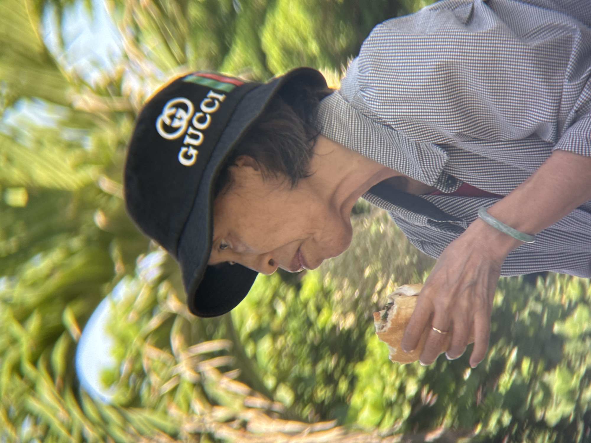 A medium shot shows an elderly Asian woman wearing a black Gucci bucket hat and a gray and white checkered jacket. She is holding a sandwich in her right hand and appears to be taking a bite. She is also wearing a thin gold ring on her left ring finger and a jade bracelet on her left wrist. The background is blurred green foliage, suggesting an outdoor setting in Nha Trang, Vietnam.