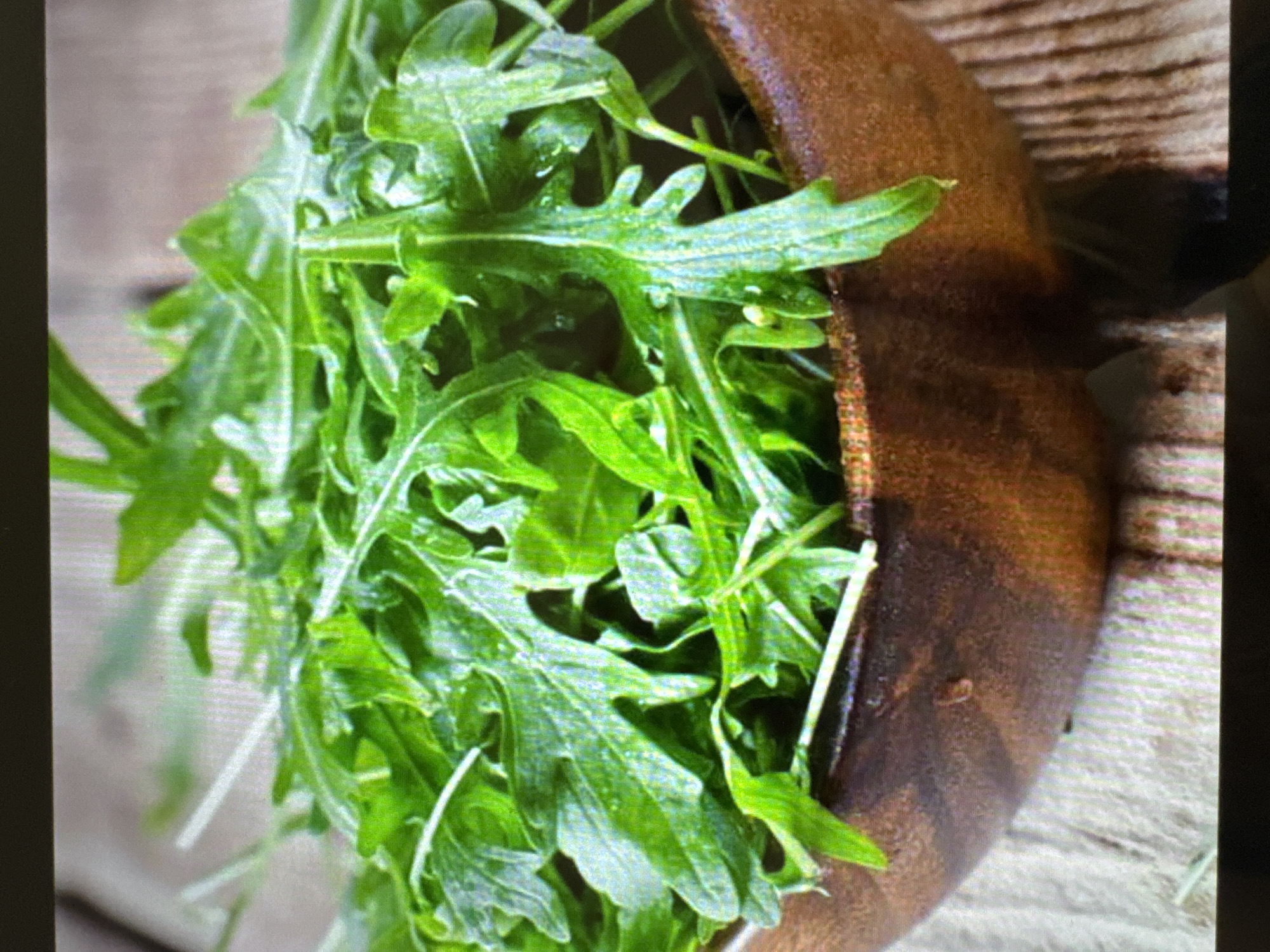 A wooden bowl overflowing with fresh arugula is placed on a rustic wooden surface. The arugula is vibrant green and appears to be recently washed, with some water droplets visible on the leaves. The bowl itself is a dark, rich wood with visible grain. The background is blurred, but it seems to be a continuation of the wooden surface. The lighting suggests an indoor setting, possibly a kitchen or dining area, with soft light illuminating the arugula. There are no people or specific landmarks visible in the image that would indicate a location in Yalta, Ukraine.