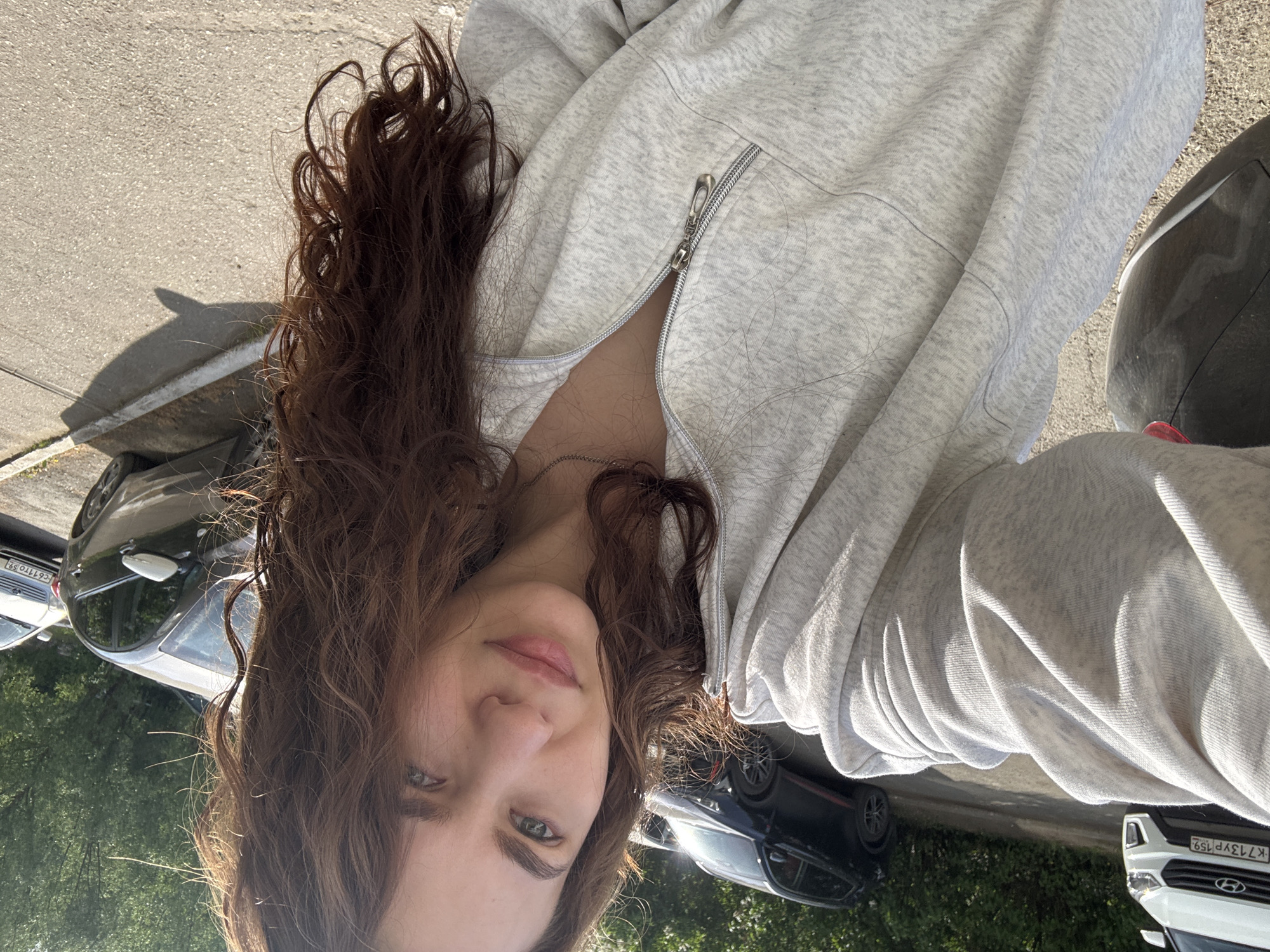 A woman with wavy brown hair and light eyes is taking a selfie outdoors on a sunny day in what appears to be an urban street in Chaikovsky, Russia. She is wearing a light gray zip-up hoodie, slightly unzipped. Behind her are several parked cars of varying colors, some greenery, and a concrete pavement. The license plate visible reads "K713YP 159" which corresponds to the Perm Krai region in Russia.