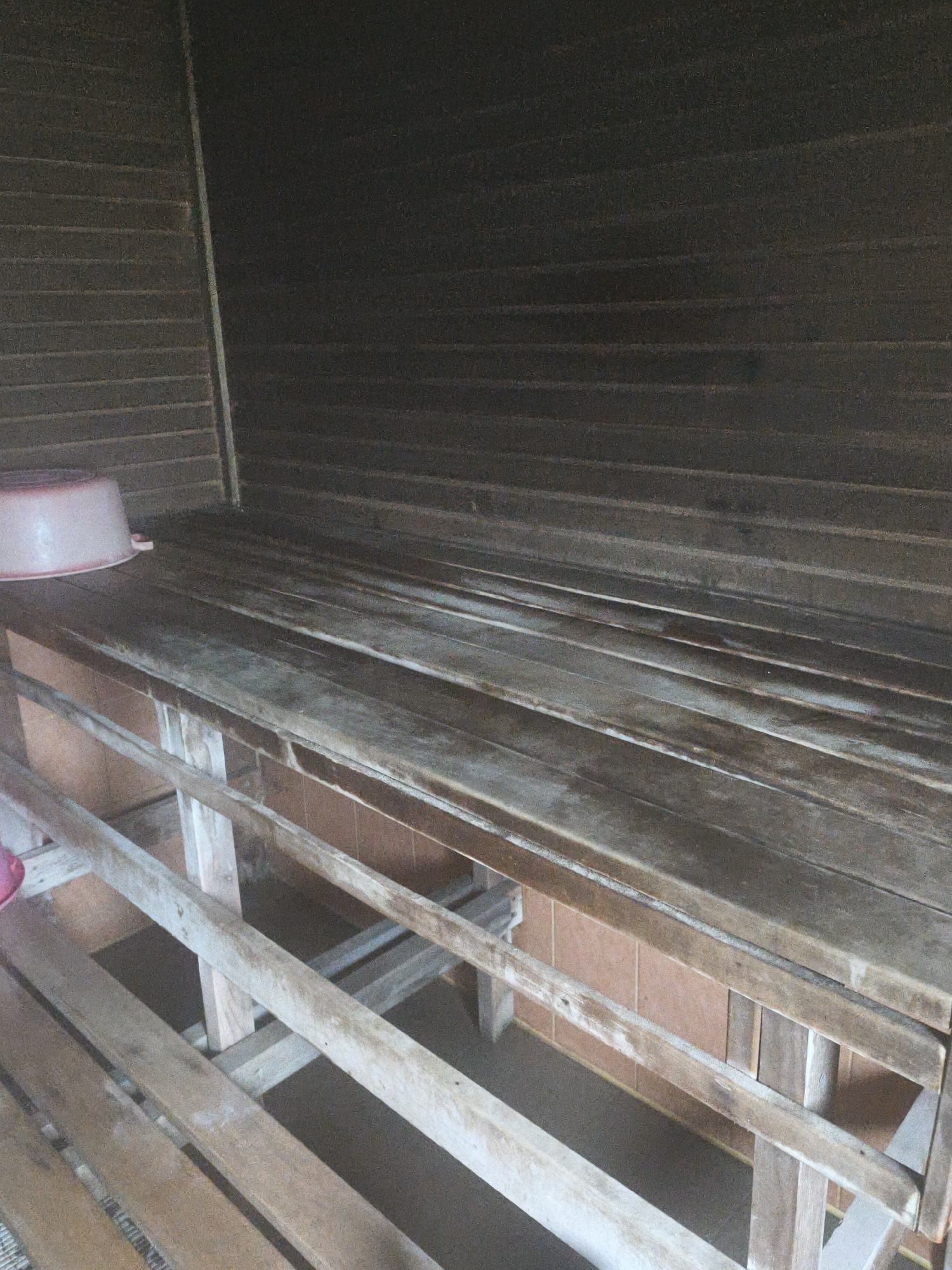 The image shows a wooden bench inside what appears to be a sauna or a similar wooden room. The bench is made of several wooden planks. In the background, the walls are also made of horizontal wooden planks. A light pinkish plastic basin is visible on the left side of the bench. The floor beneath the bench is not clearly visible, but there appears to be some tiling or a different material. The lighting is dim, suggesting an indoor environment. The location is identified as Brodi, Latvia.
