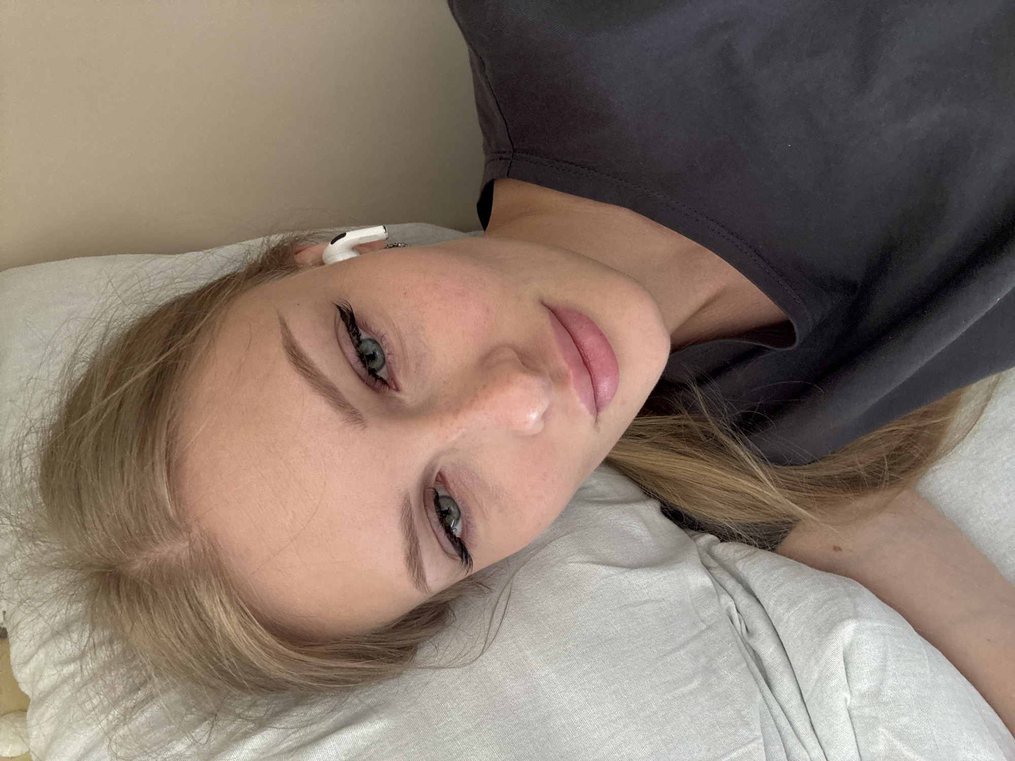 A young woman with blonde hair is lying on a bed, looking at the camera. She has on a dark gray t-shirt and is wearing white earbuds. Her left arm is bent and resting on a light-colored pillow. The background is a plain beige wall. The scene appears to be indoors. There is no information about the location in the city of Russkii Urmat, Russia.