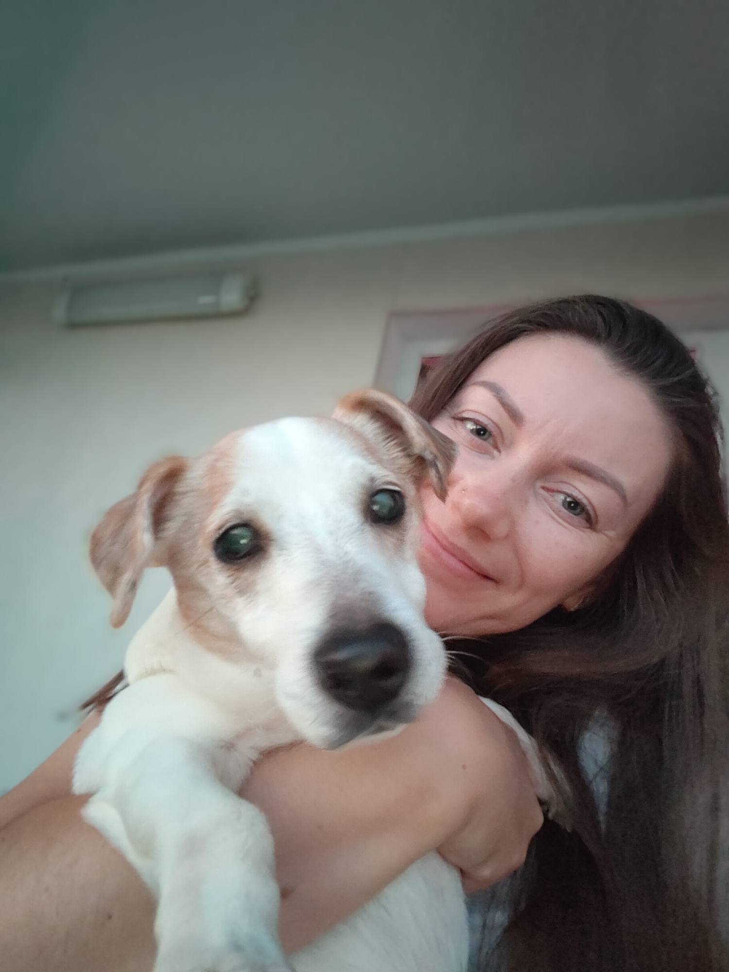 A woman with long, dark brown hair holds a small, white and tan dog in her arms. The woman is smiling at the camera, and her face is partially obscured by the dog. The dog has green eyes and its head is tilted. They are indoors, and the background appears to be a wall with a light fixture. The lighting suggests it is daytime.