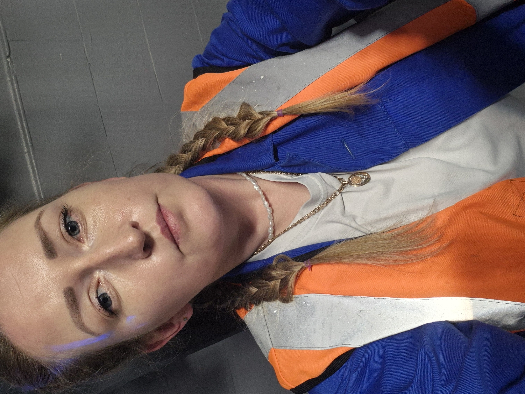 This is a portrait photograph of a young woman with blonde hair braided into a pigtail, wearing a blue and orange high-visibility jacket over a white t-shirt. She is also wearing a pearl necklace and a gold pendant. The woman has light skin, blue eyes, and appears to be wearing makeup. A faint blue line of light is visible on her cheek. The background is a plain, dark gray surface that appears to be a wall or floor. The lighting is artificial and casts some shadows. The image is oriented sideways. The photograph appears to have been taken indoors. There is no visible text in the image.