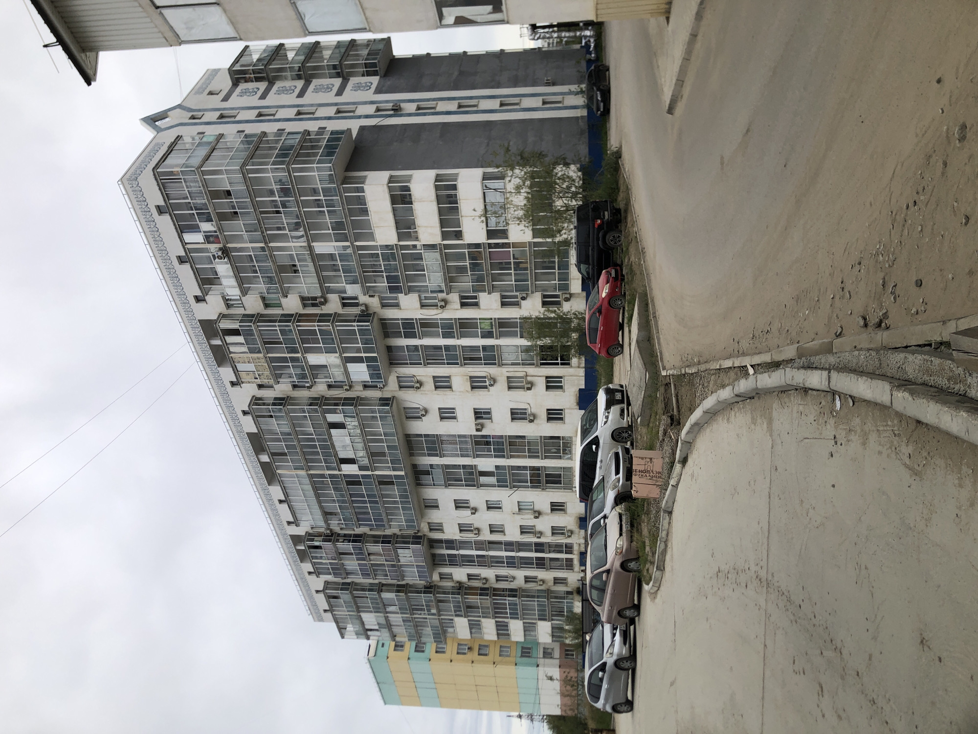 The image depicts a modern apartment building in Yakutsk, Russia. The building is tall, with multiple floors and numerous balconies, many of which are enclosed with glass. The facade is predominantly white, with grey and blue accents. In front of the building, several cars are parked along a paved road. The sky is overcast, suggesting a cloudy day. The surrounding area appears to be a residential district with other buildings and a bit of greenery.