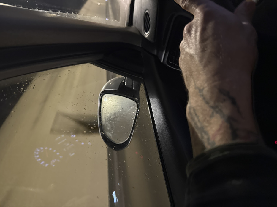 A partial view from inside a vehicle shows the passenger-side door panel, window, and exterior mirror, indicating travel within the Sozina Tunnel in Montenegro. The foreground features a person's right forearm and hand, resting on the dark interior trim. The hand displays visible veins and dark, abstract tattoos on the forearm. Raindrops cover the exterior of the passenger window and the side mirror. Through the wet window, the brightly illuminated, light-colored walls of the tunnel are visible. A reflection of the vehicle's illuminated instrument cluster, specifically a circular gauge with numeric indicators and additional digital readouts, is distinctly projected onto the window glass.