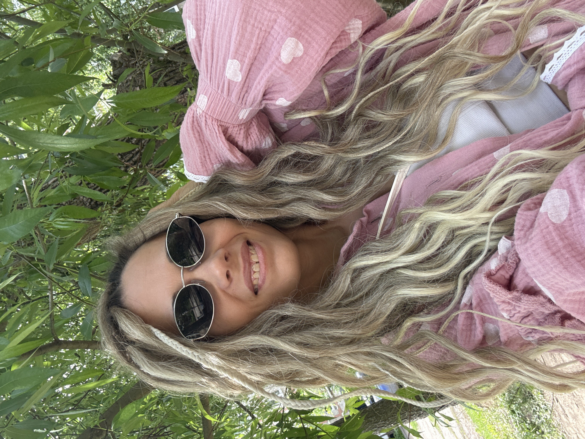 A young woman with long, flowing, wavy blonde and light brown hair is captured in a close-up, low-angle shot, outdoors in Rusanovo, Russia. Her hair, which appears to have some braided or dreadlock elements in white, is spread out around her. She wears round, dark sunglasses pushed up on her head, partially obscuring her forehead. Her face is turned slightly towards the viewer, revealing a broad, happy smile with visible teeth. She is dressed in a pink garment, possibly a top or dress, patterned with white hearts and featuring white lace trim on the collar or cuff. The fabric has a crinkled texture.
The setting is lush and green, with abundant tree leaves and branches visible in the background, suggesting a natural environment like a park or forest. A tree trunk is also partially visible amongst the foliage. The bright lighting indicates it is daytime, likely sunny. The woman appears relaxed and joyful. There is no visible text in the image.