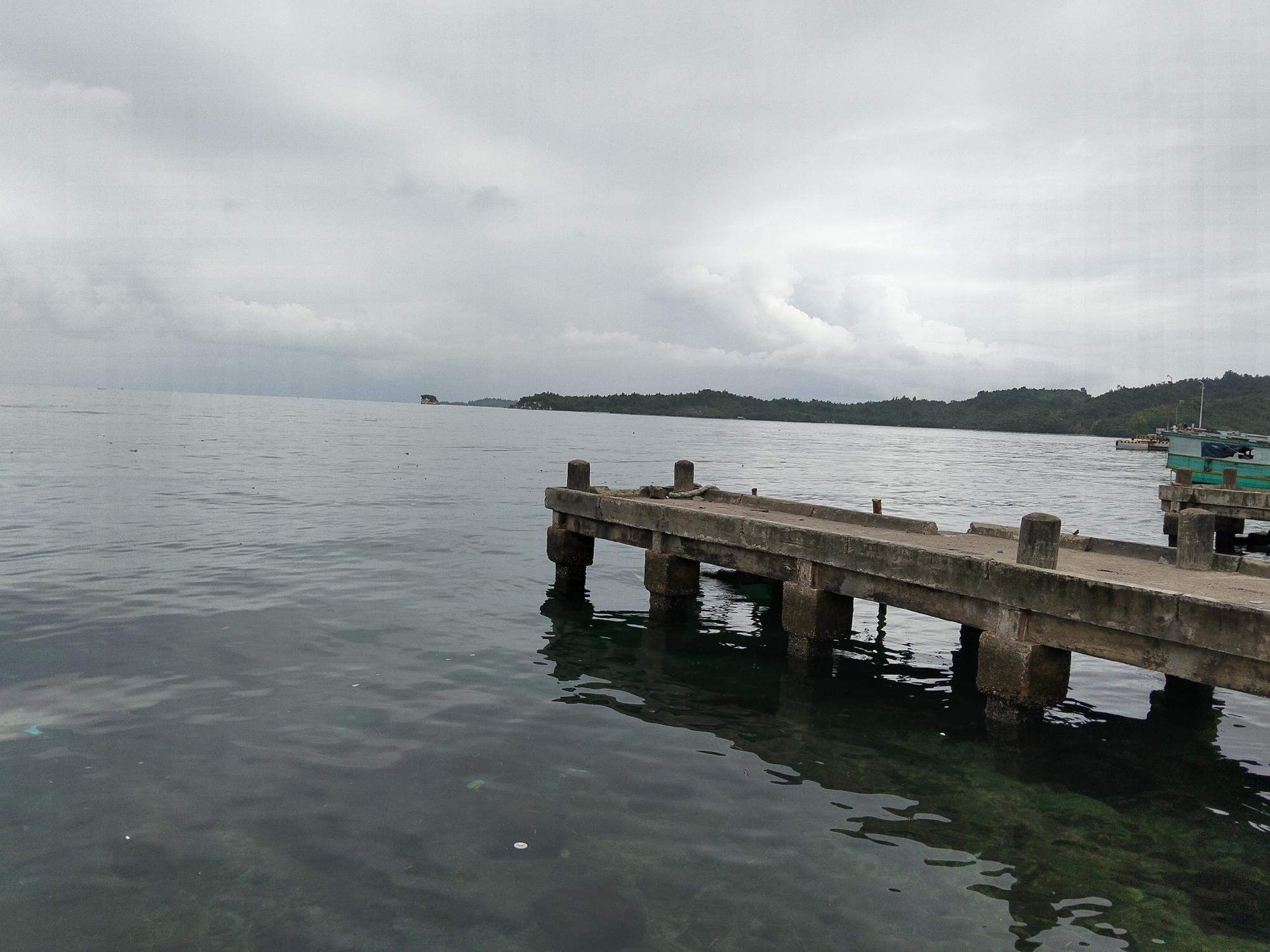 A concrete pier juts out into a calm, grey sea under an overcast sky. The pier is supported by several concrete pilings that disappear into the murky water. The surface of the pier is weathered and shows signs of age. In the distance, a lush green island or coastline is visible, shrouded in a light mist. On the right side of the image, another, smaller dock is partially visible with a boat moored to it. The water is clear enough in places to reveal the seabed below. The overall scene is one of natural beauty and a sense of quietude, possibly near Banggai, Indonesia, a region known for its coastal landscapes. There are no people visible in the image.