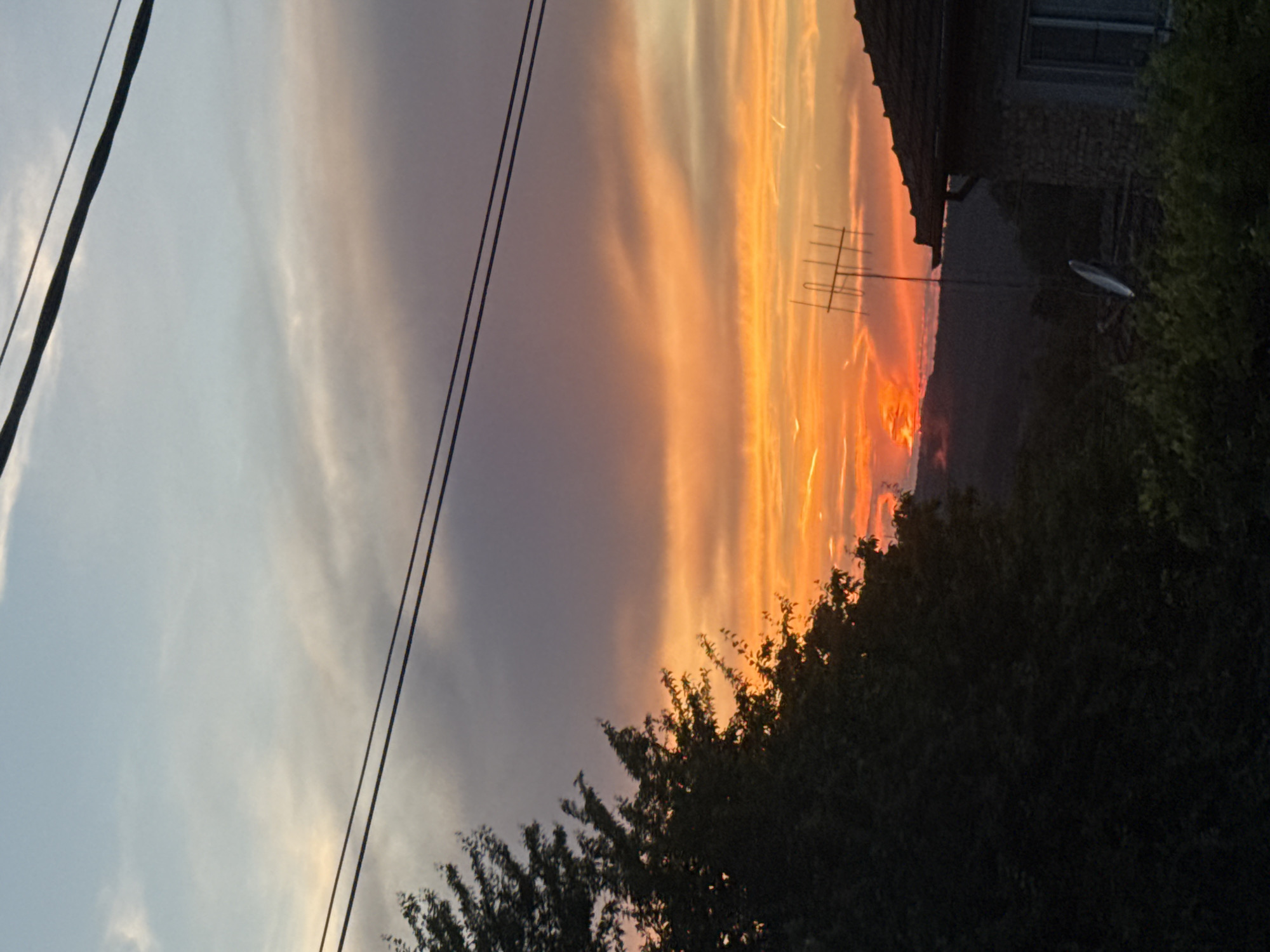 This image depicts a rural sunset scene in Krushevets, Bulgaria. The sky is filled with vibrant orange and purple hues, with wispy clouds stretching across the horizon. In the foreground, a large tree with dark foliage dominates the left side of the frame, its branches reaching towards the sky. A silhouette of a house with a tiled roof and a window is visible on the right, along with a satellite dish and an antenna. Power lines traverse the sky, adding a linear element to the composition. The background features rolling hills, shrouded in the fading light of dusk. No people are visible in the image.