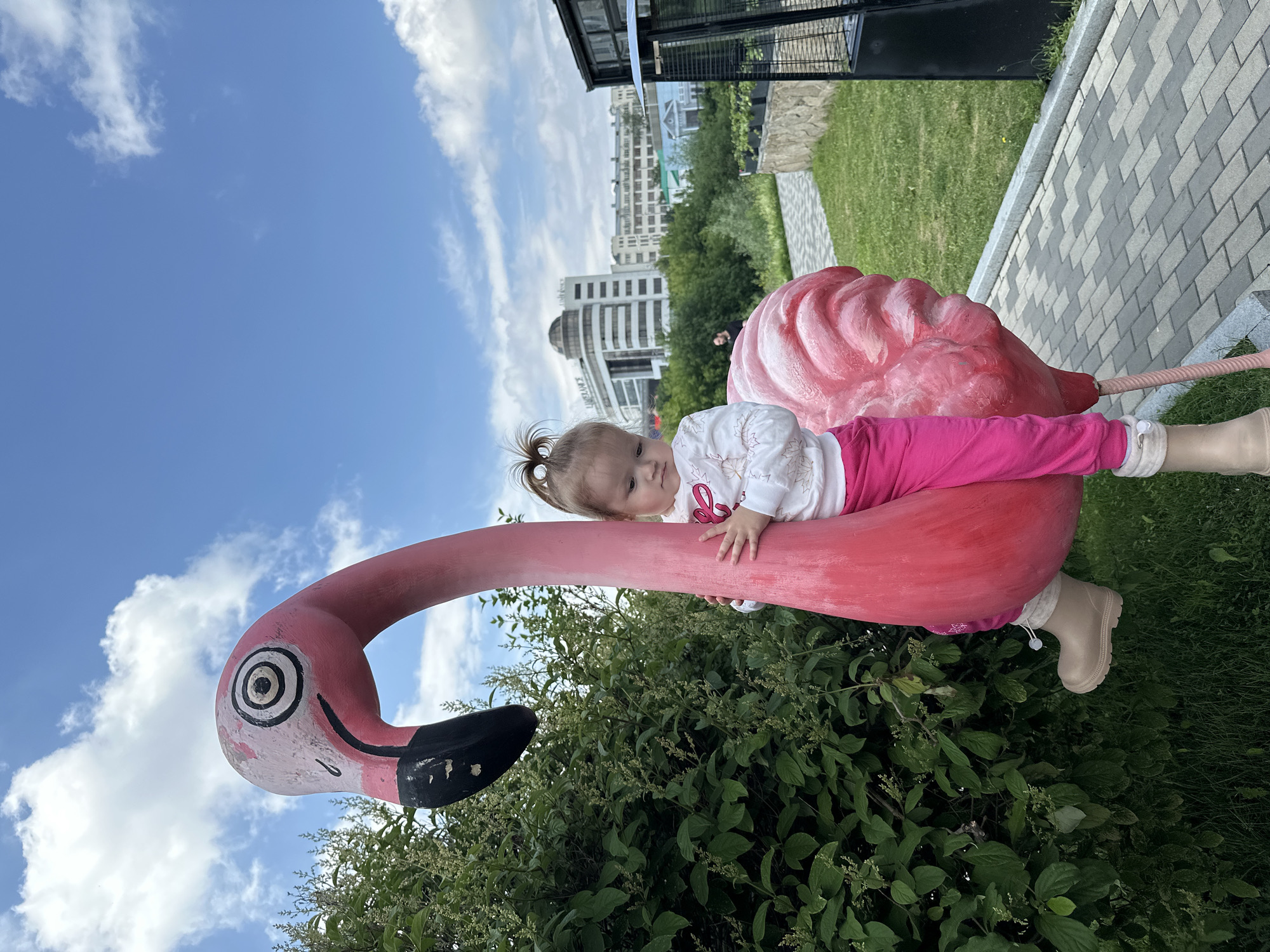 In Yekaterinburg, Russia, on a clear day with scattered clouds, a young girl with blonde hair tied in a ponytail is seated on a large, pink flamingo statue. She is wearing a white long-sleeved shirt with a pink design and bright pink pants, along with beige boots. Her hands are resting on the flamingo's neck and body. The statue is positioned outdoors, with lush green bushes to the left and a paved walkway made of gray and white bricks to the right. In the background, several modern buildings and trees are visible under the blue sky.