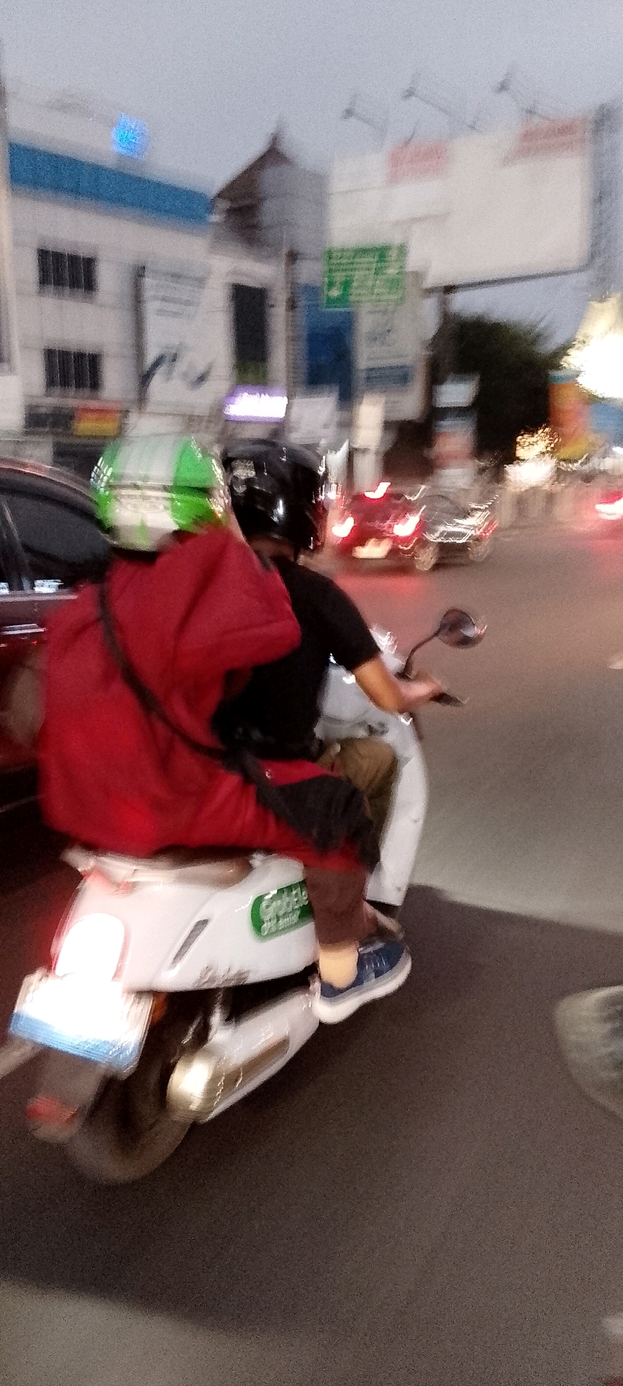 An action shot captures two individuals on a white scooter in Bekasi, Indonesia, at dusk. The person in the back, wearing a green and white helmet and a red overcoat, is blurred with motion. The driver, wearing a black helmet, is also motion-blurred, with their face obscured by the helmet. They are riding on a paved road, with buildings and streetlights visible in the background, suggesting an urban setting. The scooter has a green "GrabElectric" logo on its side, indicating it is part of a ride-sharing service. The overall image is characterized by motion blur and low light conditions, typical of a moving vehicle at twilight.
