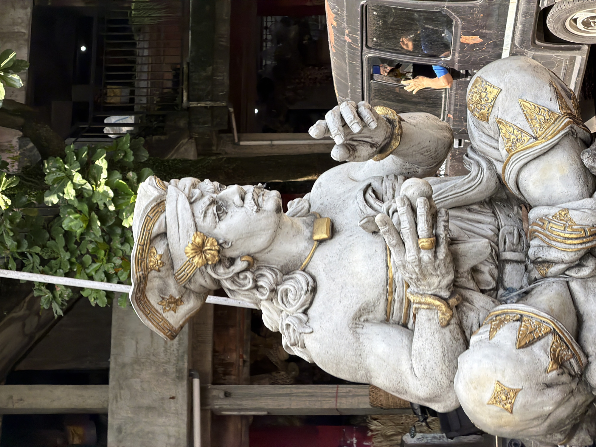 A life-size stone statue of a seated man with a stern expression and ornate headdress is the primary subject of this image, taken in the Ubud District of Indonesia. The statue is depicted in a seated position, with its legs crossed and hands held in front of its chest. It is adorned with gold accents on its headdress, jewelry, and clothing. Behind the statue, a dark-colored vehicle is partially visible, with a person's arm and hand reaching out of the window. The background consists of structures with trees and foliage, suggesting an outdoor setting, possibly a temple or a cultural site. The lighting indicates it is daytime, with shadows cast by the statue and surrounding elements.